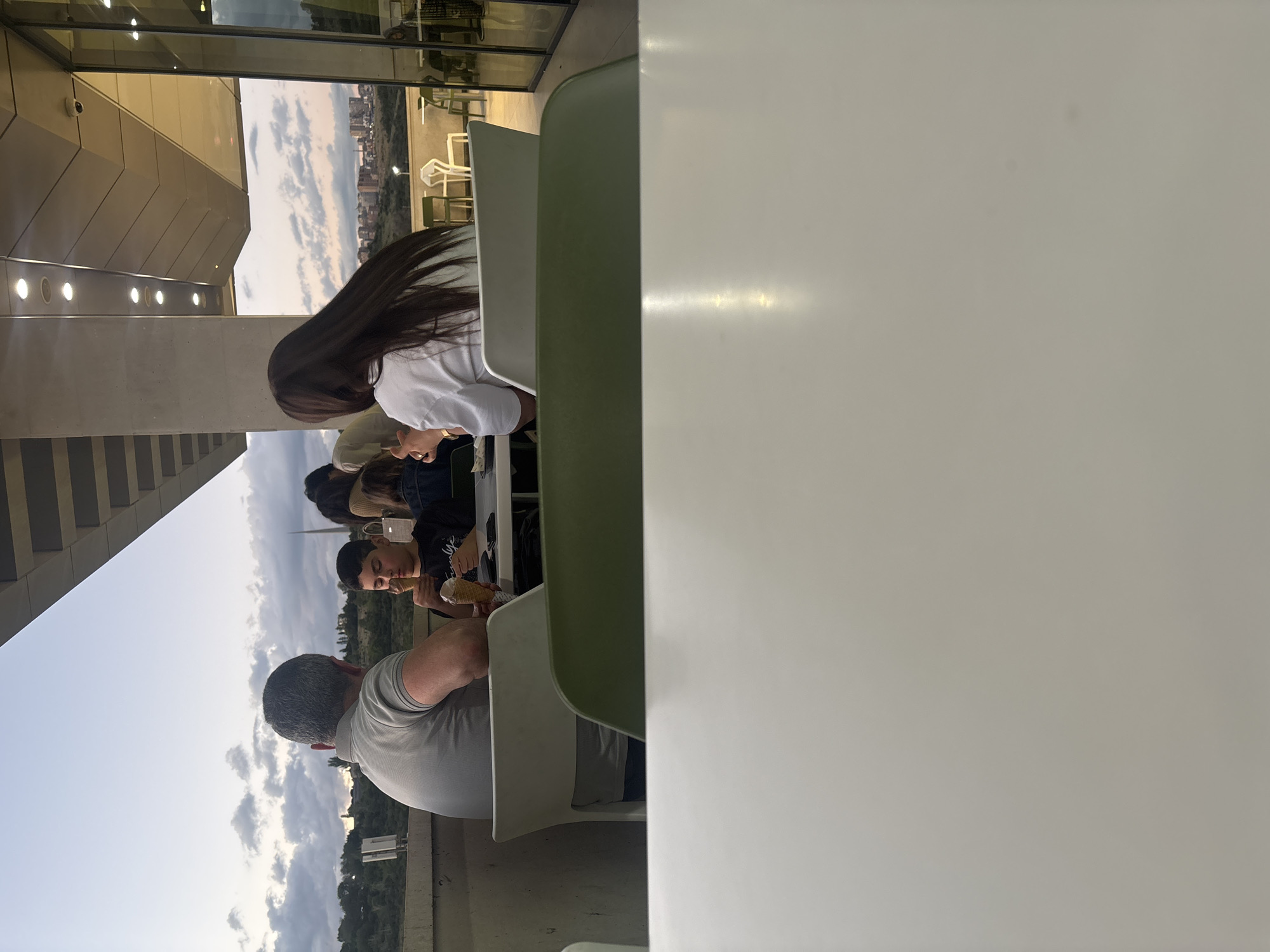 The image depicts an outdoor or semi-outdoor seating area, likely a cafe or restaurant terrace, in Yerevan, Armenia, captured during what appears to be late afternoon or early evening under a partly cloudy sky. The perspective is from a low angle, with a large, light-colored table occupying most of the right foreground.
Several individuals are seated at tables in the middle ground. Closest to the camera on the left, a person with short, grey hair and a light-colored polo shirt is seen from behind, seated at a light-colored chair. Directly in front of them, a young male with dark hair, wearing a dark shirt, is seated and appears to be eating an ice cream cone. Another person is partially visible behind him. Further left and back, a person with long, dark hair, dressed in a white long-sleeved top, is also seated, facing away from the viewer. The chairs are of a modern design, some light green and others off-white.
To the far left of the frame, a contemporary building with angular architectural features and reflective windows is visible. Circular light fixtures can be seen inside the building, and a small security camera is mounted on its exterior. Beyond the seating area, the sprawling cityscape of Yerevan extends into the distance, with buildings nestled against a backdrop of hills and a dramatic, cloudy sky. The overall atmosphere appears relaxed, suggesting a casual gathering where people are enjoying treats like ice cream. No visible text is present in the image.