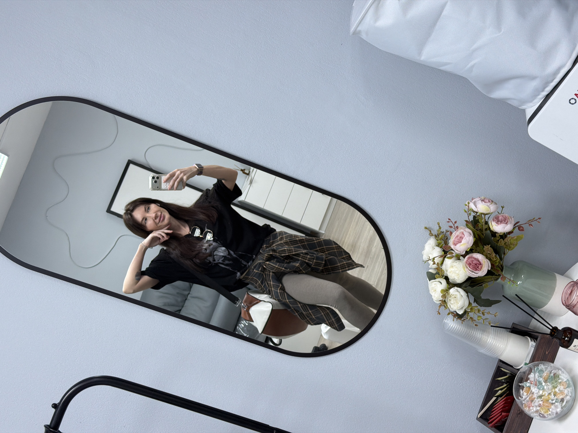The image depicts a mirror reflecting a woman seated in a chair, taking a selfie with her smartphone. She is dressed in a black T-shirt with a graphic, a plaid skirt, and light-colored tights. A purse rests on the chair next to her. The reflected background includes a gray couch and what appears to be a picture frame on the wall. The mirror itself is oblong with a black frame and is positioned against a light blue wall. To the right of the mirror, on what is presumably a table or surface, there is a bouquet of artificial flowers in a vase, a stack of plastic cups, an aroma diffuser, and a decorative bowl filled with colorful candies or objects. Part of a black metal frame or fixture is visible at the bottom left of the image. The location is identified as Perm, Russia.