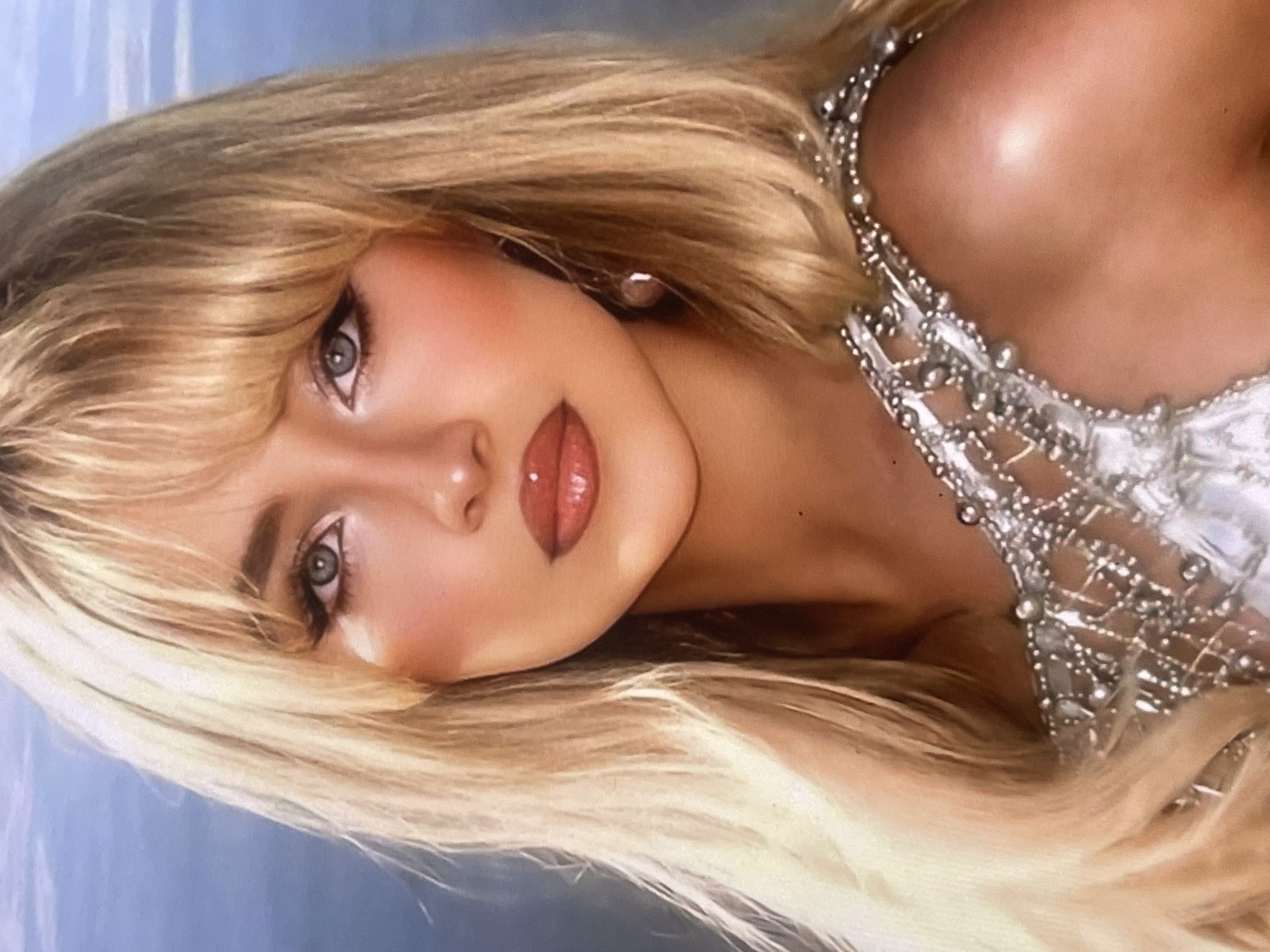 The image features a portrait of a woman with long, voluminous blonde hair, parted with bangs that frame her face. Her head is tilted slightly, and her gaze is directed towards the viewer with a calm and confident expression. Her eyes are light-colored, possibly blue or grey, accentuated with dark eyeliner and mascara. She has defined eyebrows, subtle blush on her cheeks, and full, glossy lips in a reddish-brown or nude tone.
She is wearing an elaborate garment, likely a dress or top, that is light-colored (possibly white or silver) and heavily embellished with beading, sequins, or small pearls, particularly visible on the shoulder and upper chest area. A small, light-colored earring is visible on her right ear. The texture of the embellishments suggests a delicate, intricate design.
The background is a softly blurred, light blue tone, suggesting a plain backdrop or a distant sky, which provides a gentle contrast to her features and attire. The lighting is soft, highlighting her facial features and the sheen of her hair and makeup. The overall impression is one of elegance and glamour.
No specific activity beyond posing for a portrait is evident. There are no visible location cues within the image itself to suggest Poltava, Ukraine, other than the provided context. No text is visible in the image.