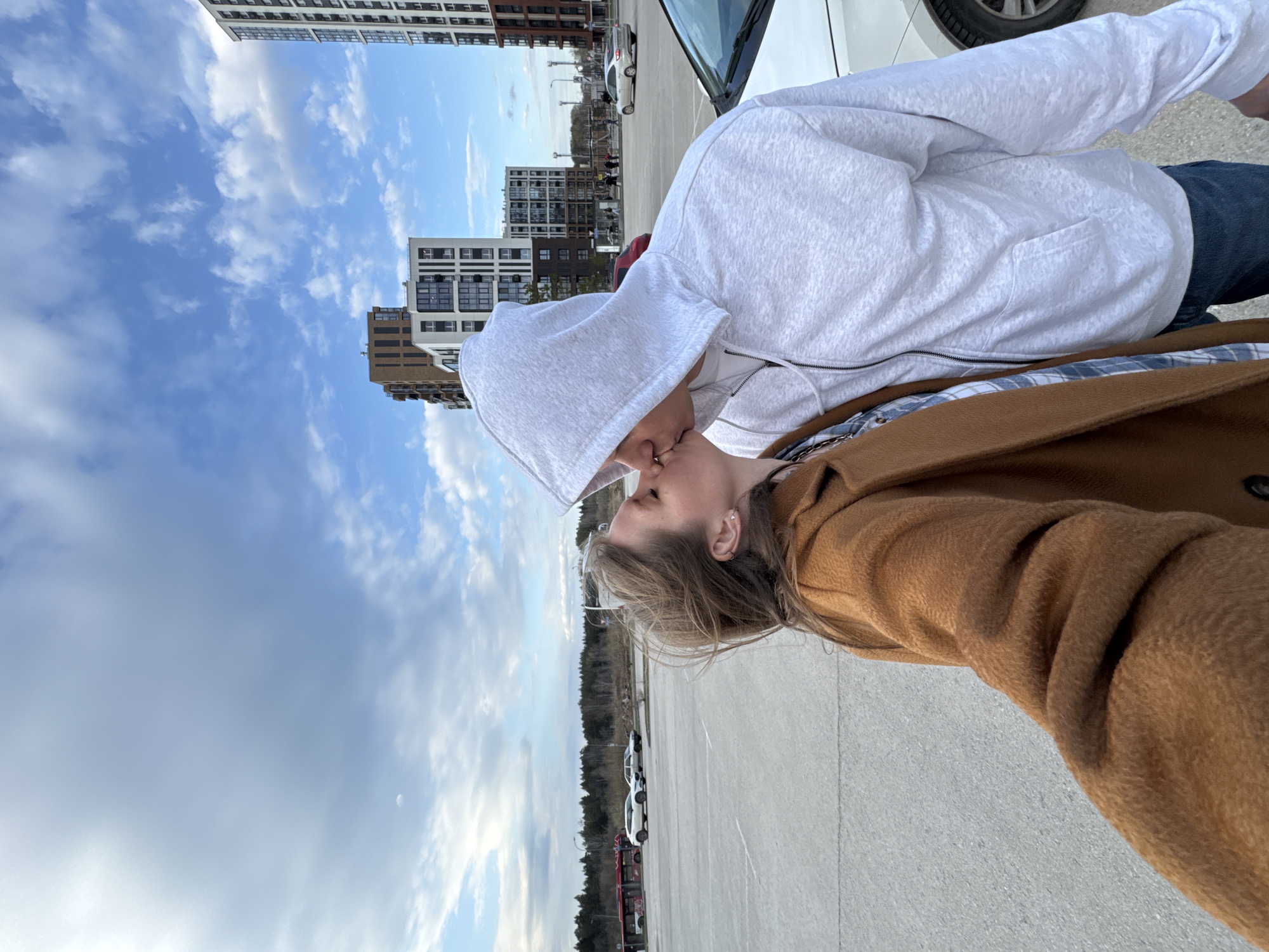 The image captures a close-up, intimate moment between two individuals kissing outdoors. The photograph is oriented on its side, but when viewed upright, it reveals a horizontal scene.
On the left, a person wearing a light grey zippered hoodie with the hood up is seen leaning in for the kiss. Their face is partially obscured by the hood as their lips meet the other person's. They also wear blue jeans. On the right, the second person wears a warm brown coat over a blue and white plaid shirt. Their long, light brown hair cascades down, and a visible ear sports small earrings. The embrace suggests a moment of affection, possibly captured as a selfie.
The setting appears to be an urban or suburban environment. In the background, under a partly cloudy blue sky, stand several modern, multi-story apartment or office buildings. The ground is a light grey paved surface, likely a parking lot. A white car is partially visible behind the couple, and other vehicles are parked further in the distance. Beyond the urban structures, a line of trees or a wooded area is visible on the horizon.
The weather is fair, with bright daylight indicating either late morning or afternoon. The sky shows a mix of white cumulus clouds and clear blue patches. The overall mood is tender and romantic. This scene, featuring modern high-rises and a parking lot, is characteristic of contemporary developments found in cities like Perm, Russia. No visible text is present in the image.