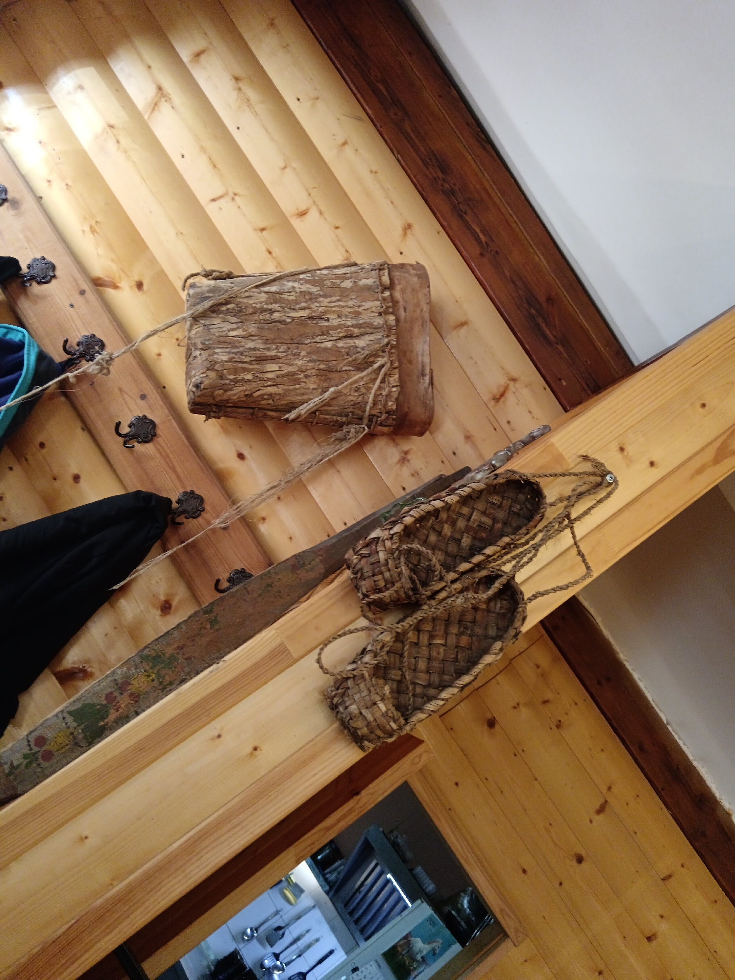 The image displays a rustic, wooden interior setting, likely in a building in Gorokhovets, Russia. The primary subjects are a woven bark container and a pair of woven shoes, possibly bast shoes (lapti). These items are suspended or displayed on wooden planks and beams. The bark container, appearing aged and somewhat rough, is secured with rope. The bast shoes are also made of woven material, with rope straps.
The background consists of wooden paneling and structural beams, suggesting a traditional or rustic architectural style. There are also several dark, ornate hooks mounted on the wooden planks, one of which is holding a black garment. To the left, a portion of a blue and black bag is visible. Below the main display, through an opening, a glimpse of kitchen utensils and shelving can be seen, indicating a functional space. The lighting appears to be natural, casting shadows and highlighting the textures of the wood and woven materials. The overall impression is of a display of traditional crafts or historical items within a well-preserved or recreated rustic environment.