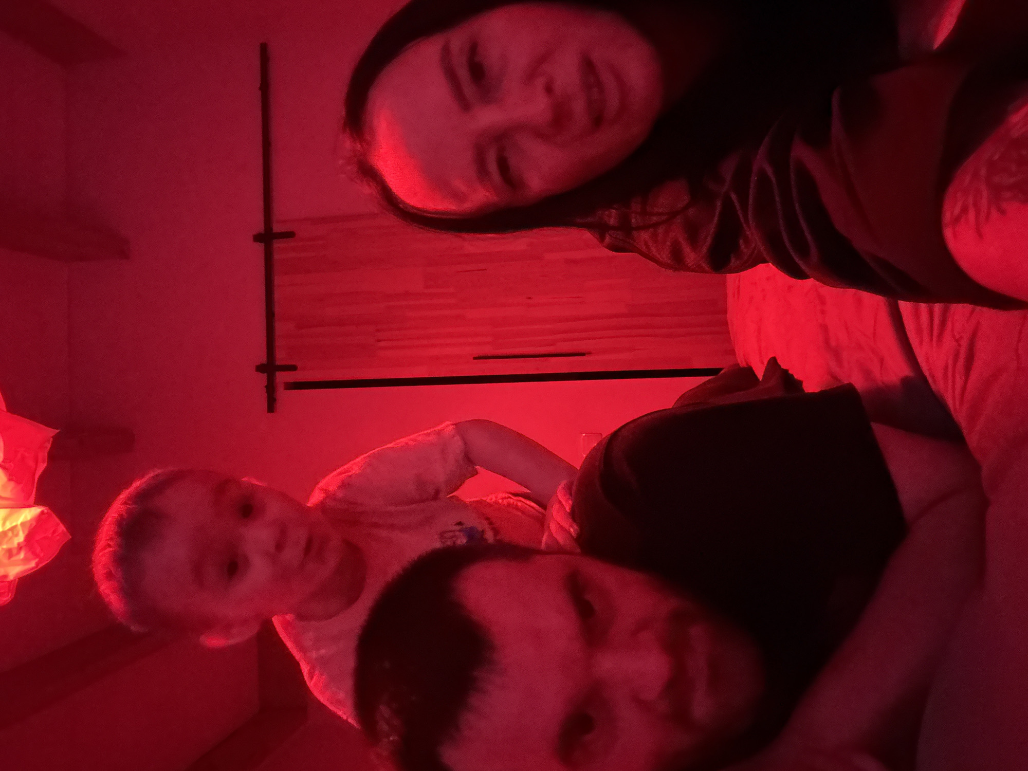 An indoor scene, illuminated by strong red artificial lighting, features three individuals in a casual, playful moment. At the top of the frame, positioned upside down, is a woman with dark hair, smiling or smirking directly at the camera. She wears a dark top, and a tattoo is visible on her right arm or shoulder. In the foreground, a man with dark hair looks towards the camera. Between the man and the inverted woman, a young child with light hair also looks towards the lens, wearing a light-colored top, with a neutral or curious expression.
The three individuals appear to be relaxing together, likely on a bed or soft surface. The tilted camera angle and inverted orientation of the woman suggest an informal, possibly selfie-style, photograph. On the far left, a decorative light fixture, resembling a paper lantern, emits a warm, albeit red-tinted, glow. Behind the woman, a wooden wall decoration or shelving unit with vertical and horizontal elements is visible. The heavy red lighting makes it impossible to discern natural colors or the exact time of day, but it implies an evening or nighttime setting. The overall mood is relaxed and intimate. There is no visible text in the image. The scene is set in Chaikovsky, Russia.