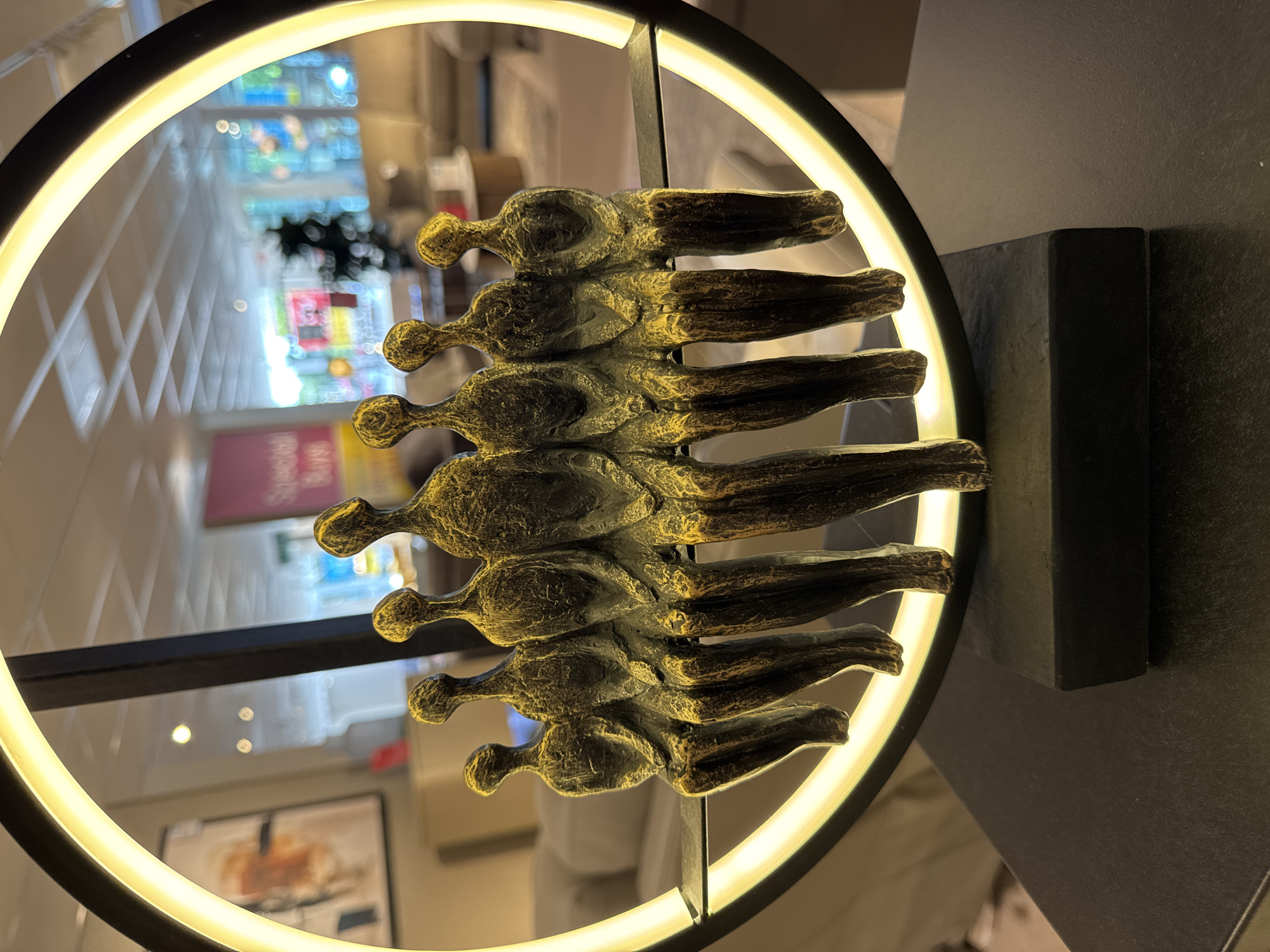 The image shows a close-up of a sculpture featuring seven abstract, elongated figures seated in a row. The figures are metallic, with a distressed gold and dark brown patina, and appear to be made of a cast material. They are seated on a dark, flat surface that is part of a larger circular object. This circular object has a bright, illuminated band around its inner edge, suggesting it is a light fixture. The sculpture is placed on a dark, rectangular base. The background is out of focus but appears to be an interior setting, possibly a furniture store or showroom, with displays of furniture, artwork, and ceiling lights visible. There are no people or animals actively interacting with the sculpture in the foreground. The location is not specified to be in London, United Kingdom.