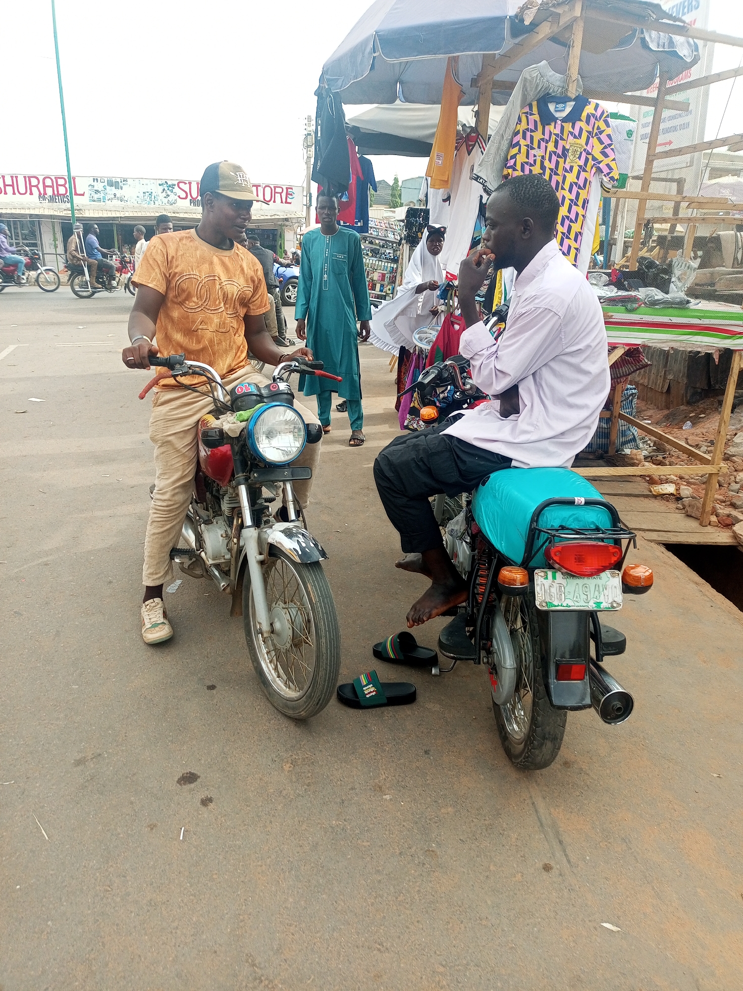 This outdoor image captures a lively street scene in Tempure, Nigeria, likely during daylight hours. The setting appears to be a marketplace or commercial area, with various shops and stalls.
In the foreground, two men on motorcycles are central to the composition, seemingly engaged in conversation. The man on the left wears an orange t-shirt with an "AUDI" logo, tan pants, and a dark cap. He is seated on a red and silver motorcycle, resting his left foot on the ground, wearing white sneakers. The man on the right, facing towards the left, wears a light pink long-sleeved shirt and dark trousers. He is barefoot, sitting on a motorcycle with a bright blue seat cover, with black sandals placed on the ground beside his vehicle.
Behind these two, several other people are visible. To the far right, part of a market stall is seen, constructed with wood and covered by a light-colored umbrella. Various clothes, including a distinctive yellow and pink patterned jersey, hang from the stall. Further back, a man in a teal-colored long garment (agbada) stands, observing the scene. On the left side of the street, other individuals ride motorcycles, and a building with shopfronts lines the background.
Visible text includes a sign on the building in the background, partially reading "SHURABL COSME" (likely "SHURABLE COSMETICS") and "SU STORE" with "PHO" below. The license plate on the rear motorcycle clearly shows "NIGERIA" and "GATEWAY STATE" (identifying Ogun State), with the alphanumeric code "TGG-49AWG". The ground is paved, showing some dirt and a discarded cigarette butt. The overall atmosphere suggests a typical bustling day in a local commercial area.