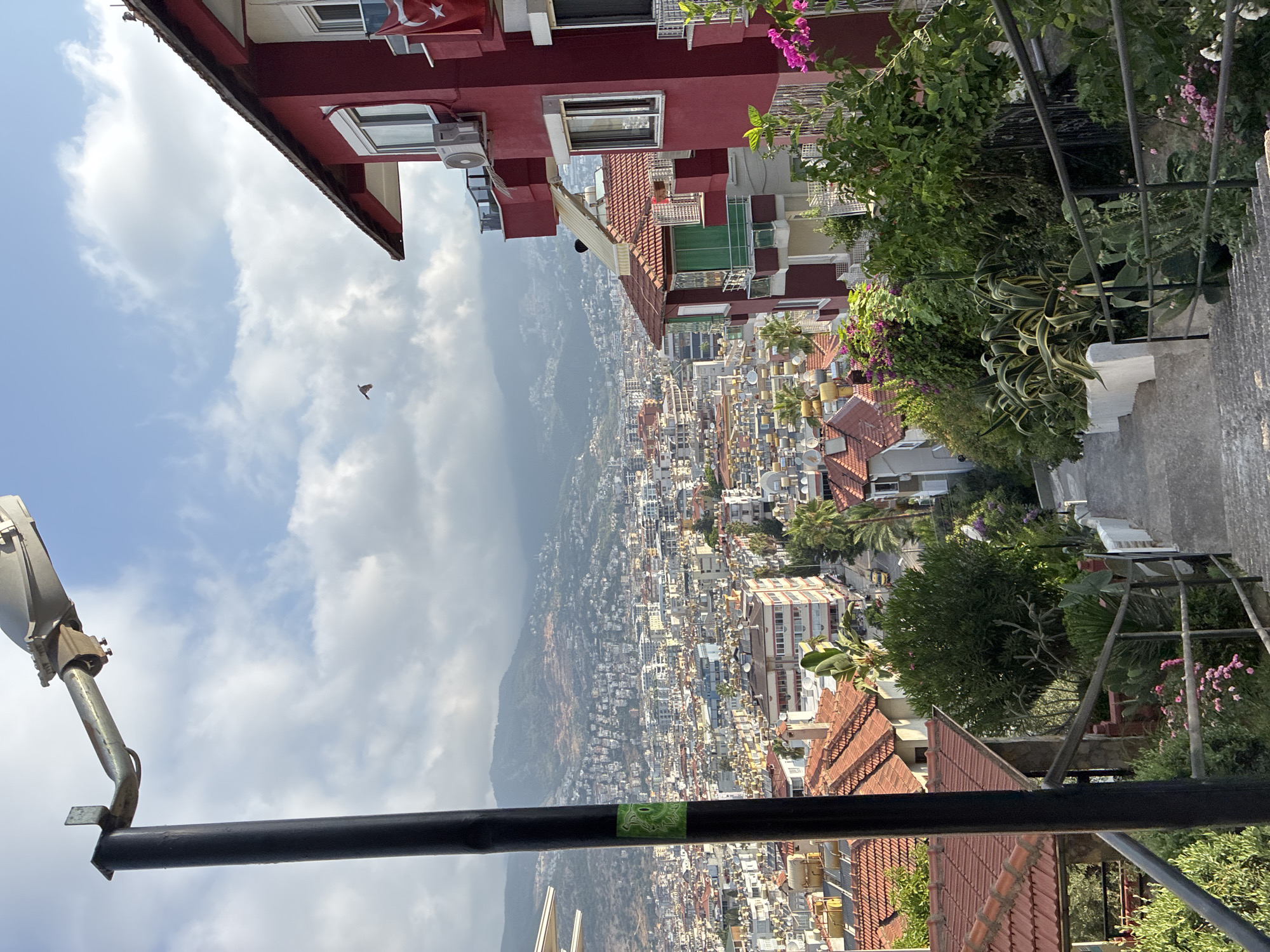The image captures a downward view of a steep, stone staircase in Alanya, Turkey, with lush greenery and flowering plants lining its sides. In the foreground, a black lamppost stands prominently to the left, partially obscuring the view. The staircase leads down towards a densely populated urban landscape stretching into the distance, characterized by a multitude of buildings with red-tiled roofs and the majestic backdrop of hazy mountains. A distinctive red building with white trim and balconies stands to the right, adorned with an air conditioning unit and a Turkish flag. The sky above is partly cloudy, with a small bird visible in flight. The overall scene depicts a residential area on a hillside overlooking the city and its surrounding natural landscape.