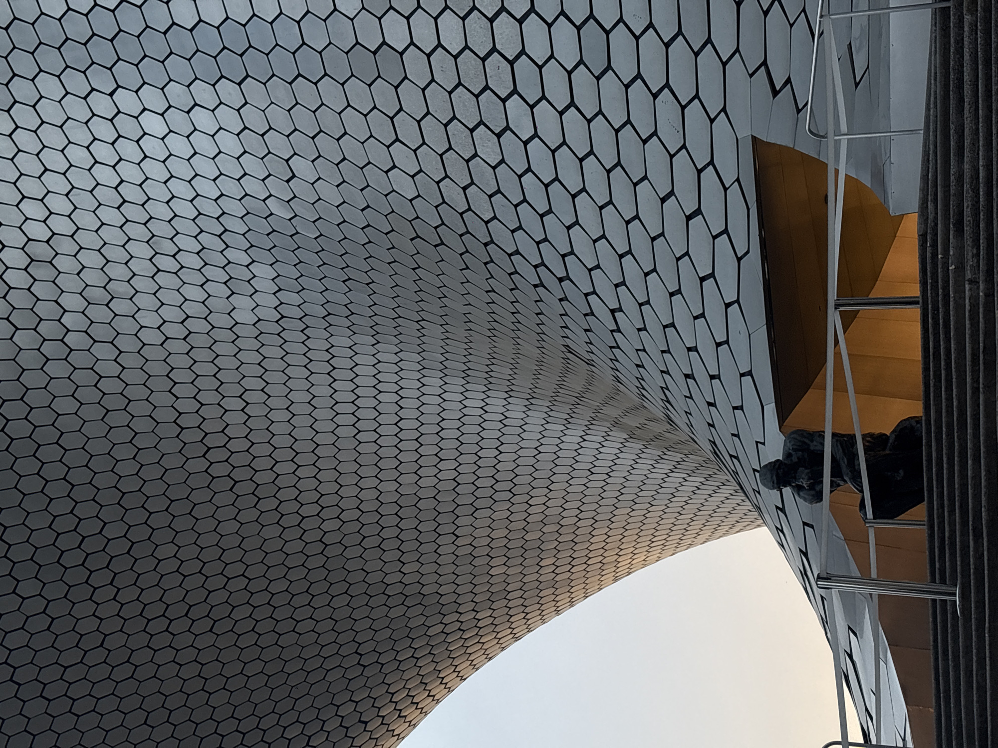 The image depicts a low-angle view of the exterior of the Museo Soumaya in Mexico City, Mexico. The museum's distinctive facade is composed of numerous hexagonal tiles. A ladder is visible along the edge of the building. Part of a metallic statue is also visible on the ladder. The sky is visible in the background.