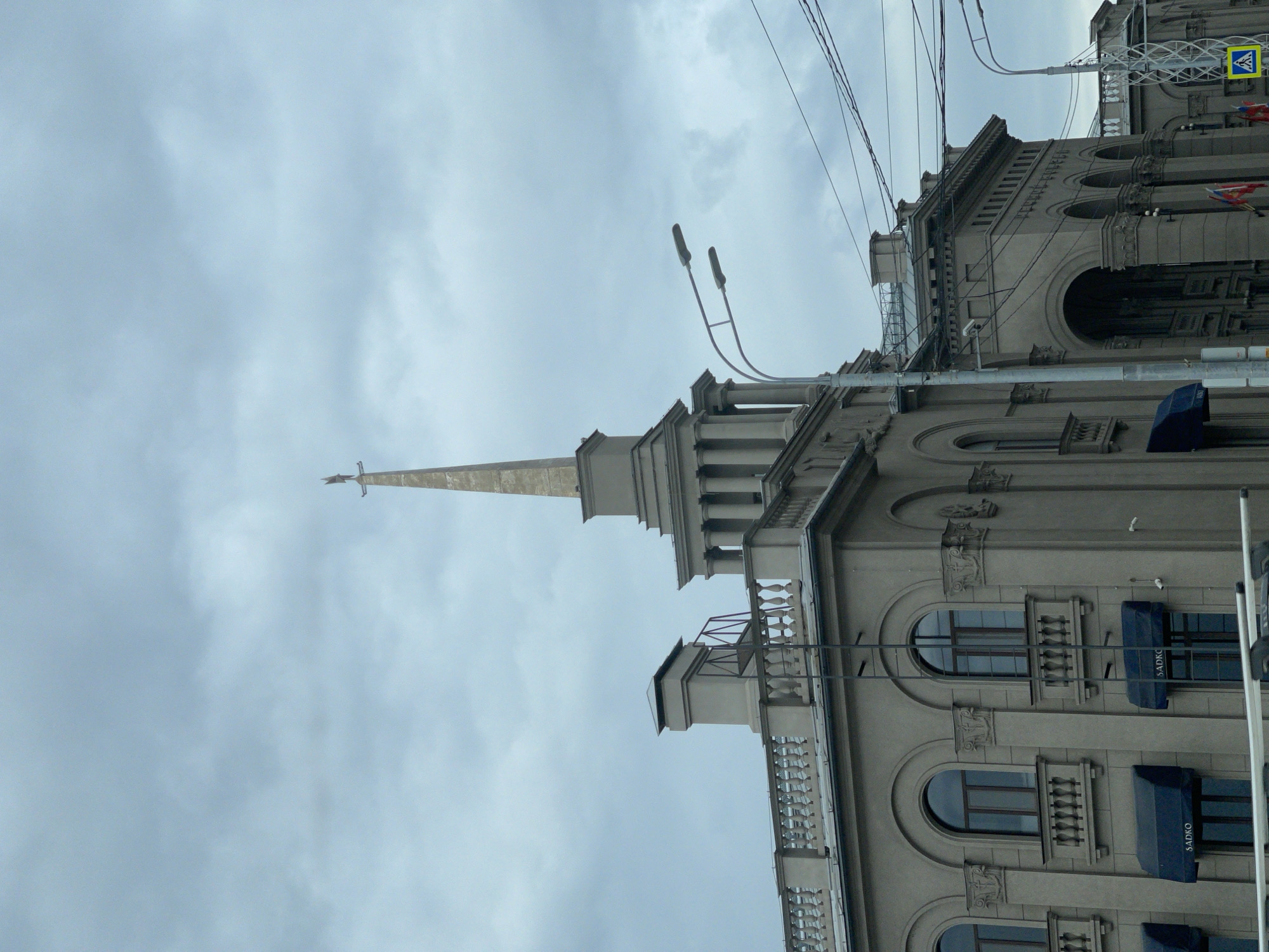 The image depicts a building in Krasnoyarsk, Russia, with a prominent spire reaching towards a cloudy sky. The building's architecture is characterized by a neoclassical style, featuring multiple stories, arched windows, ornate facades, and a visible balcony. Streetlights and power lines intersect the scene, indicating an urban setting. A sign can be seen on the building, and awnings are visible above the windows.