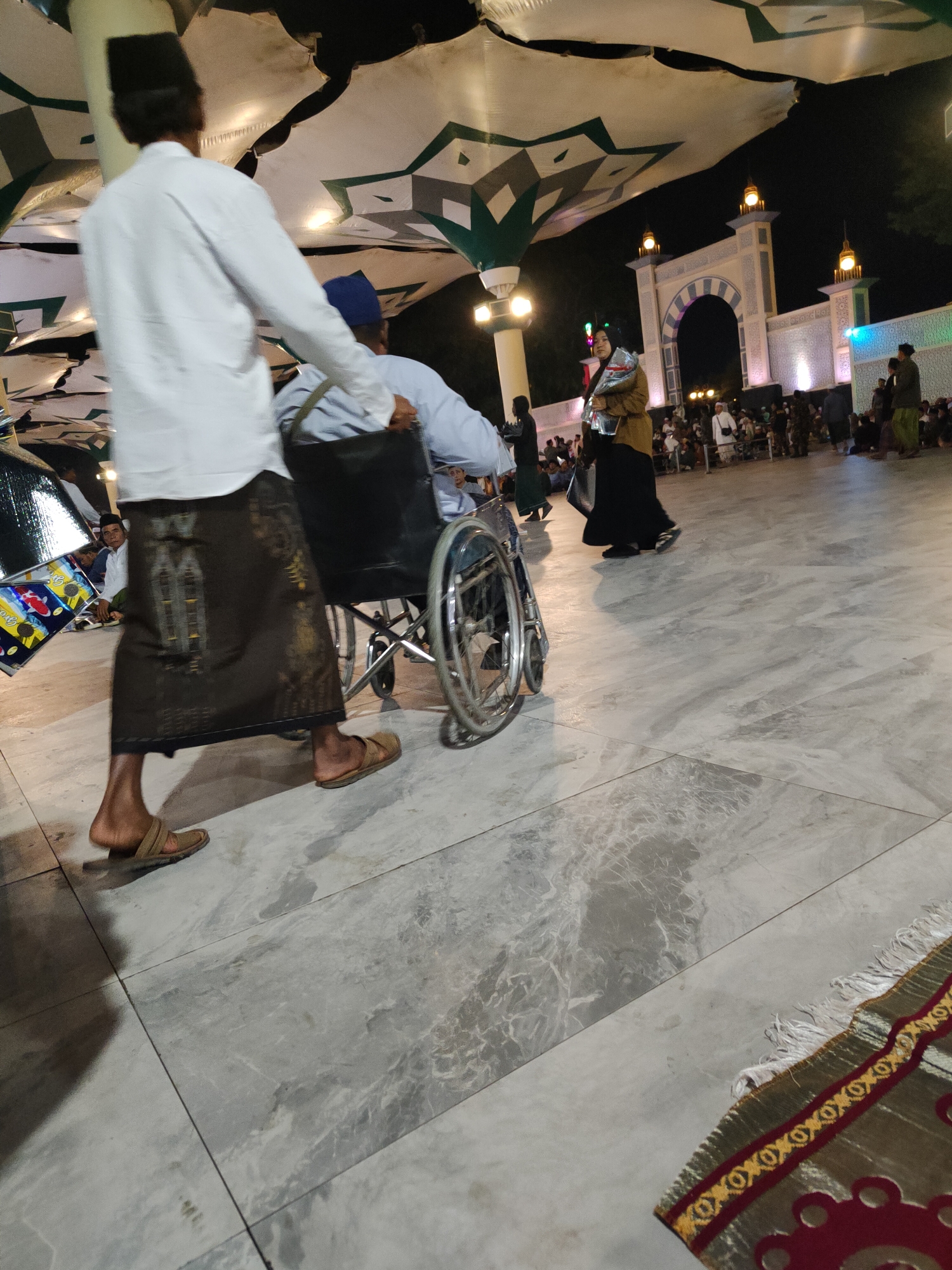 In Pasuruan, Indonesia, at nighttime, an individual wearing a white long-sleeved shirt and a dark sarong is pushing a person in a wheelchair. The person in the wheelchair is wearing a light blue long-sleeved shirt and a blue cap. Both individuals appear to be men. In the background, a woman in a brown jacket and black skirt walks away from the camera, while a mosque-like structure with illuminated arches and minarets is visible. Other people are scattered in the background, some sitting and some standing. The ground is paved with large, light-colored marble tiles. A portion of a prayer rug with a red and gold patterned border is visible in the lower right corner of the image. The scene is illuminated by artificial lights.