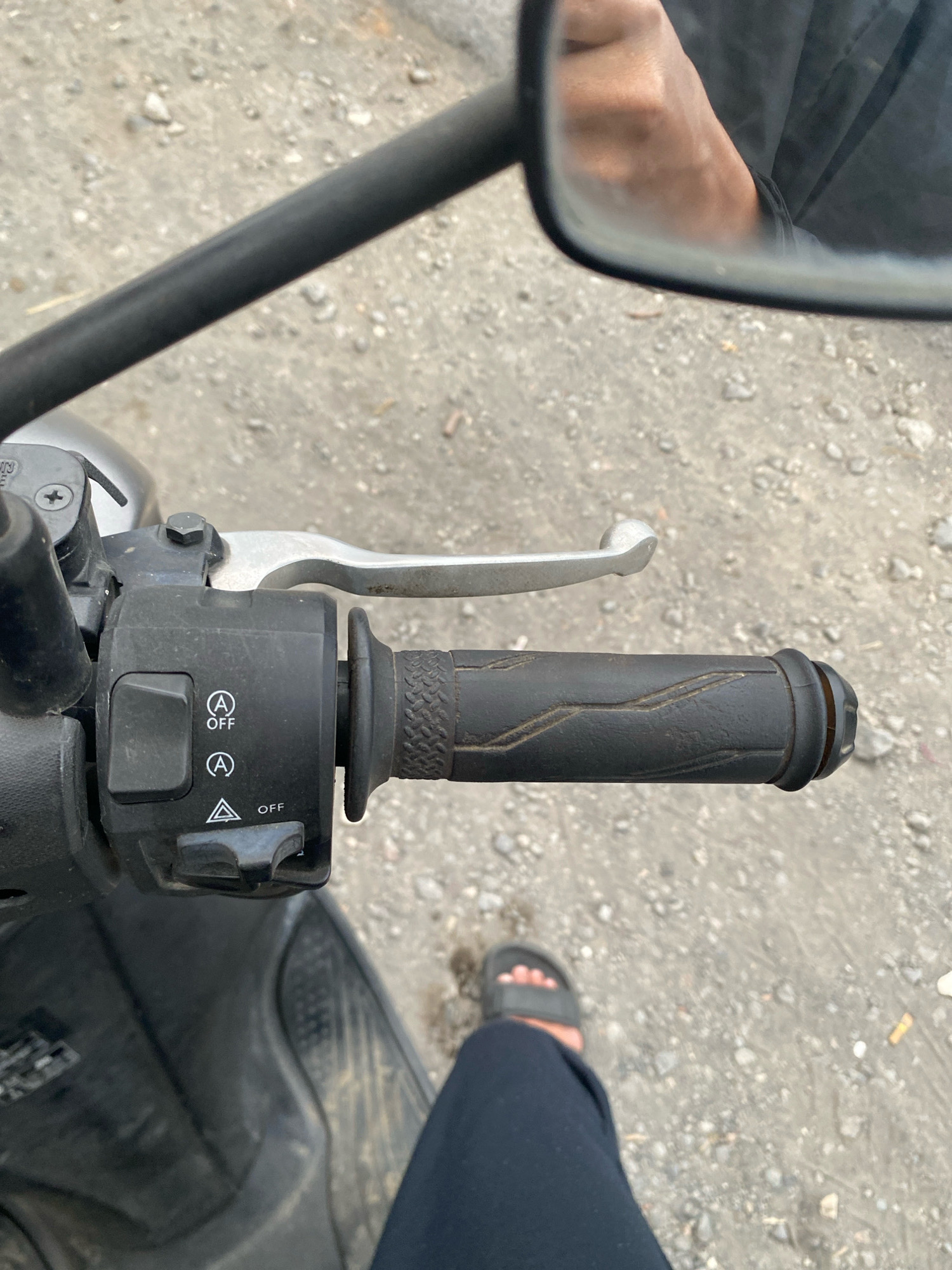 The image captures a close-up view of a motorcycle's handlebar and controls, situated on a gravelly surface in Karangluhur, Indonesia. The handlebar features a silver brake lever, a black grip with textured lines, and a black control housing. This housing contains several buttons and switches, including a square button, two circular buttons with a symbol resembling a capital "A" and the word "OFF" underneath, and a triangular symbol with the word "OFF" below it, controlled by a toggle switch. A side mirror is also visible, reflecting a person's hand and dark clothing, hinting at the rider's presence. The ground is composed of small stones and dirt, typical of outdoor or unpaved areas. The lighting suggests it is daytime.