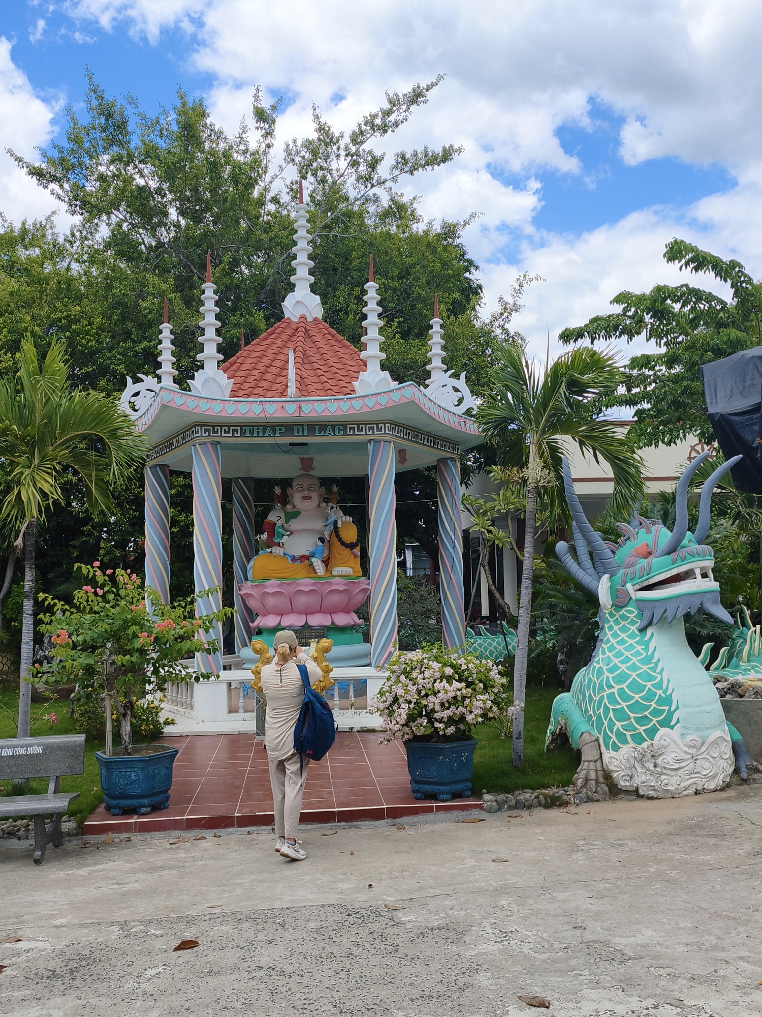 In Cam Ranh, Vietnam, a person stands in front of a colorful Buddhist shrine. The shrine features a large, multi-armed statue of a seated Buddha, adorned with gold accents and seated on a pink lotus. The shrine itself is a multi-tiered structure with a red roof and pastel-colored pillars. To the right of the shrine is a large, green dragon statue, a common symbol in Vietnamese culture. The person is wearing a light-colored outfit and a cap, and is holding a phone up to take a photo. They are also carrying a blue backpack. The ground is paved with concrete and red tiles. Lush green trees and a bright blue sky with white clouds form the backdrop. Several potted plants, including a palm tree and a flowering bush, are also visible. A stone bench is situated to the left of the frame, bearing an inscription. The scene appears to be a place of worship or a cultural attraction.
