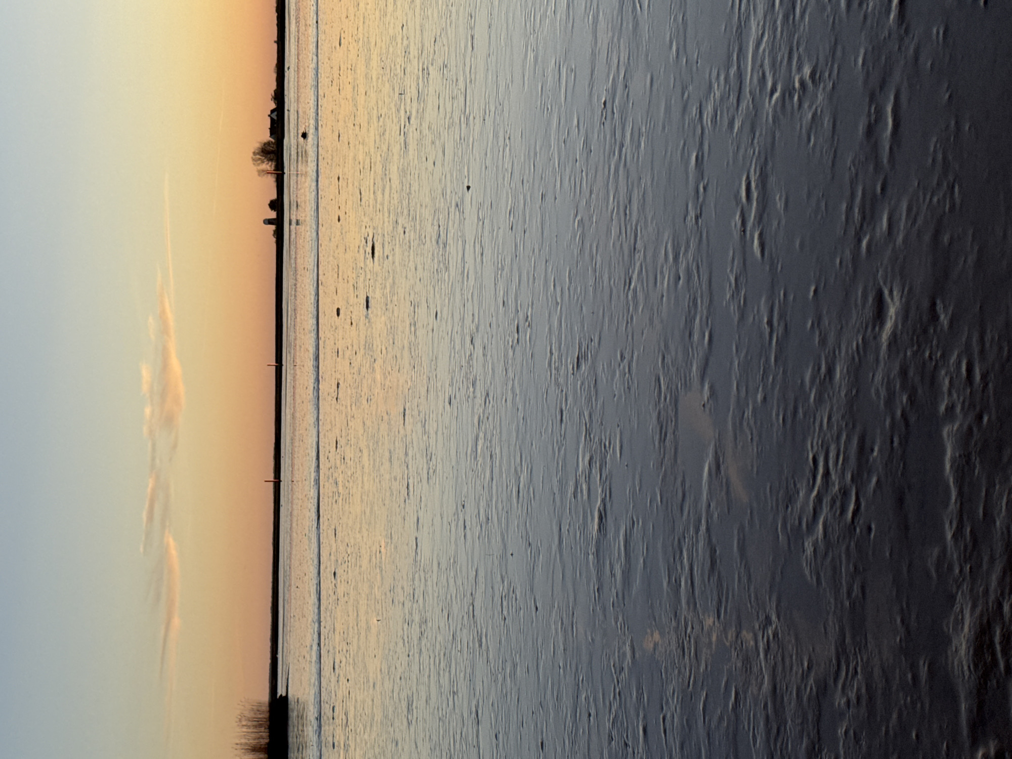The image shows a panoramic view of a shoreline at sunset. The sky transitions from a pale blue to a golden-orange hue, with wispy clouds near the left. A thin strip of land with trees and structures lines the horizon. The water reflects the colors of the sky, transitioning from a bright, reflective surface near the horizon to a dark, textured foreground. The right side of the image features muddy or sandy flats with visible patterns and shadows. The location is likely in or near Great Yarmouth, United Kingdom, due to the coastal environment and structures along the horizon. No people or specific actions are visible.