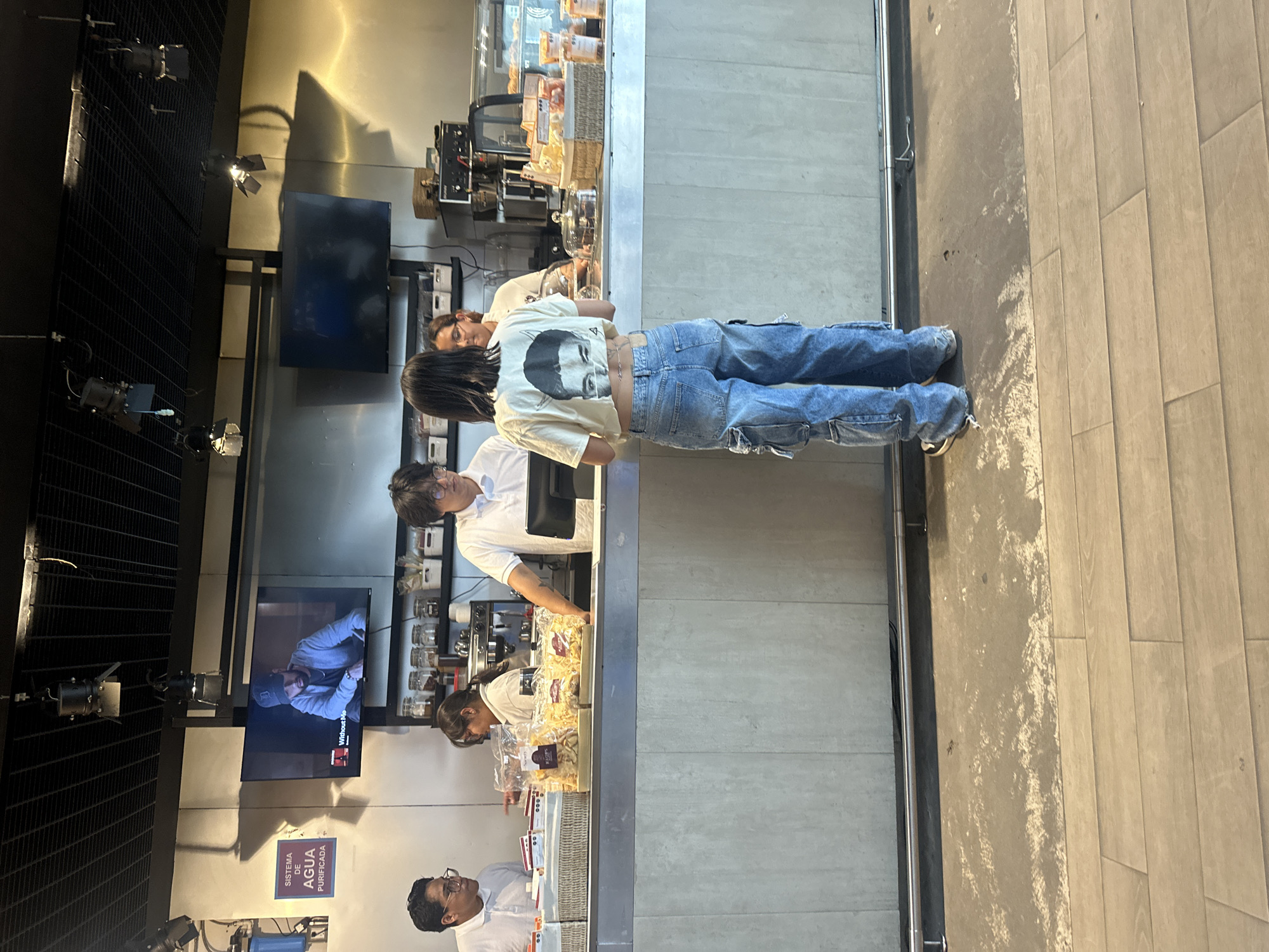 The image shows an indoor scene, likely a cafeteria or deli in Mexico City, Mexico. Several individuals are present, seemingly interacting at a counter displaying packaged goods.
A person in a t-shirt and jeans is standing at the counter, potentially a customer. Staff members in white shirts are behind the counter, possibly serving or assisting customers. One staff member is wearing glasses.
Behind the counter, shelves hold various products, and food display cases contain pastries. A television screen on the wall displays an image. The environment appears well-lit, with lights mounted on a dark ceiling structure. Signage, including a banner for "AGUA PURIFICADA" is visible. The floor is tiled.