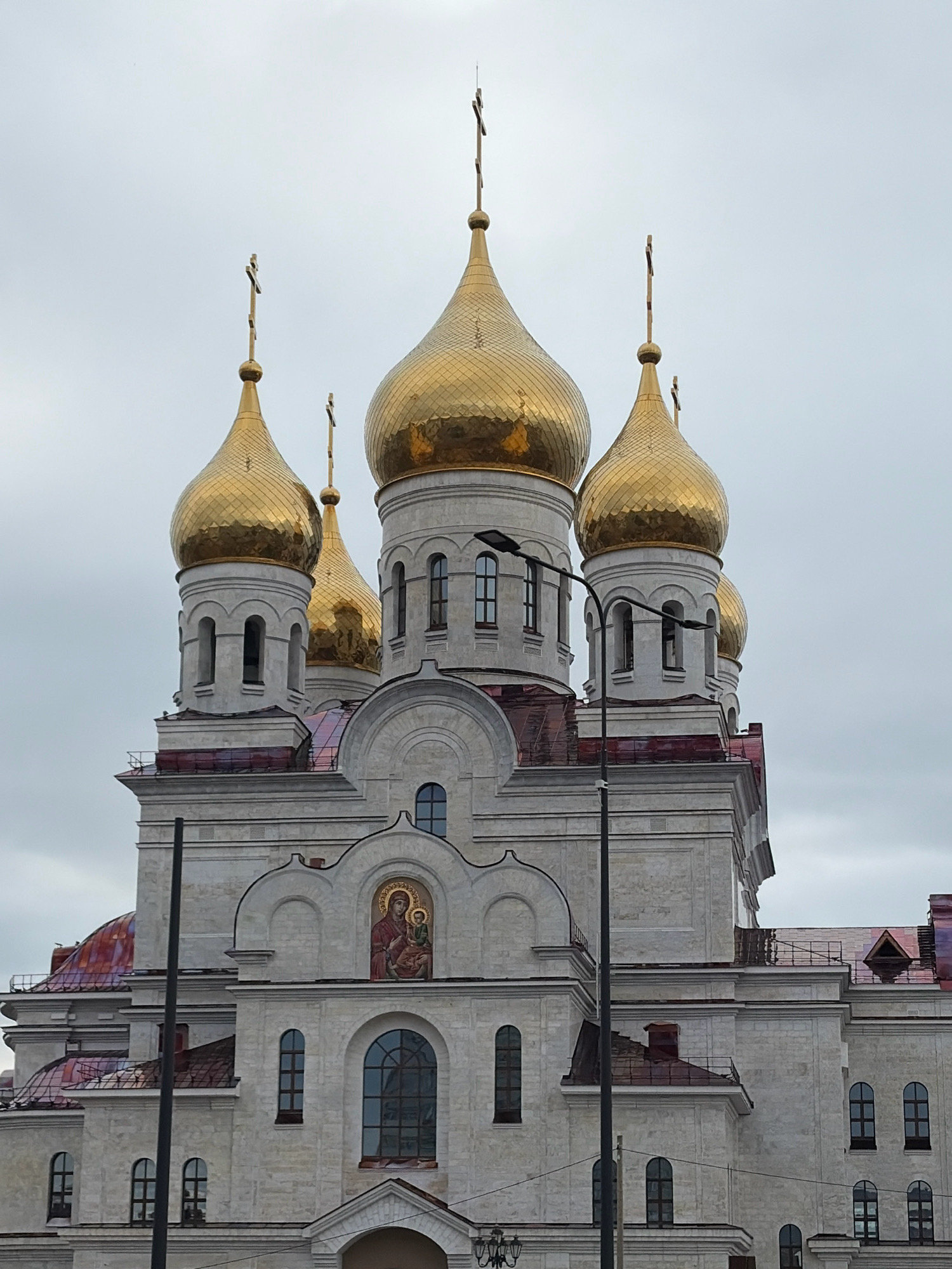 The image shows a large, ornate church with golden domes and a light-colored stone facade. The church has multiple levels and is decorated with architectural details, including arched windows and a mosaic of a religious figure. There are several golden crosses atop the domes. The sky is overcast and gray. The church is situated in a city environment, with streetlights visible in the foreground. The architecture suggests it is likely an Orthodox Christian church. This church is likely the Church of the Assumption of the Virgin Mary in Arkhangelsk, Russia, which is known for its distinctive golden domes and prominent location in the city.