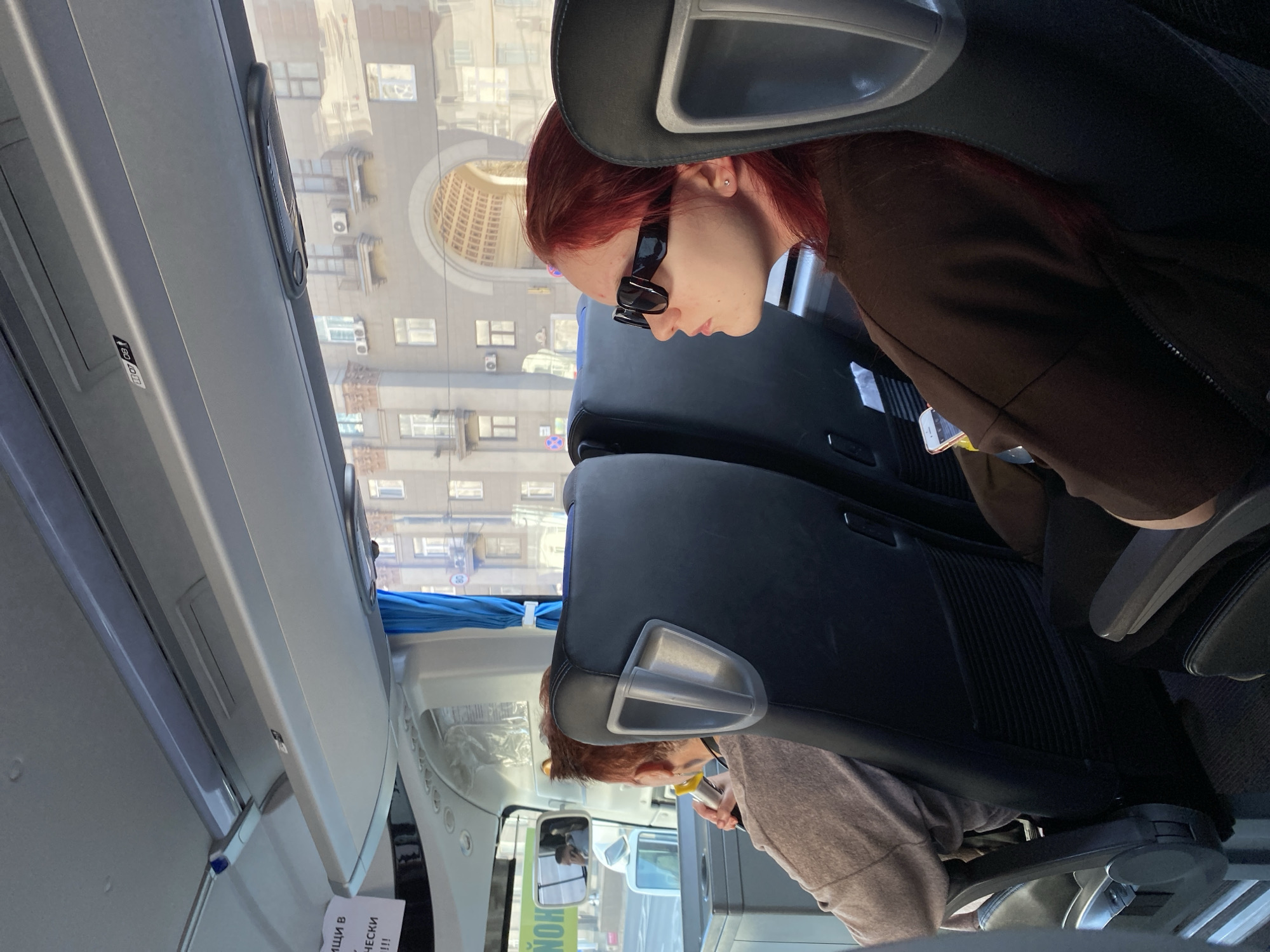 The image shows the interior of a passenger vehicle, likely a bus, with two passengers visible in the foreground. One passenger, with reddish hair and wearing sunglasses, is seated and appears to be looking at a mobile phone. A second passenger is seated behind them. Outside the window, a view of Moscow, Russia, can be seen. The architecture suggests an urban environment, with a large building featuring an arched structure visible in the reflection of the window.