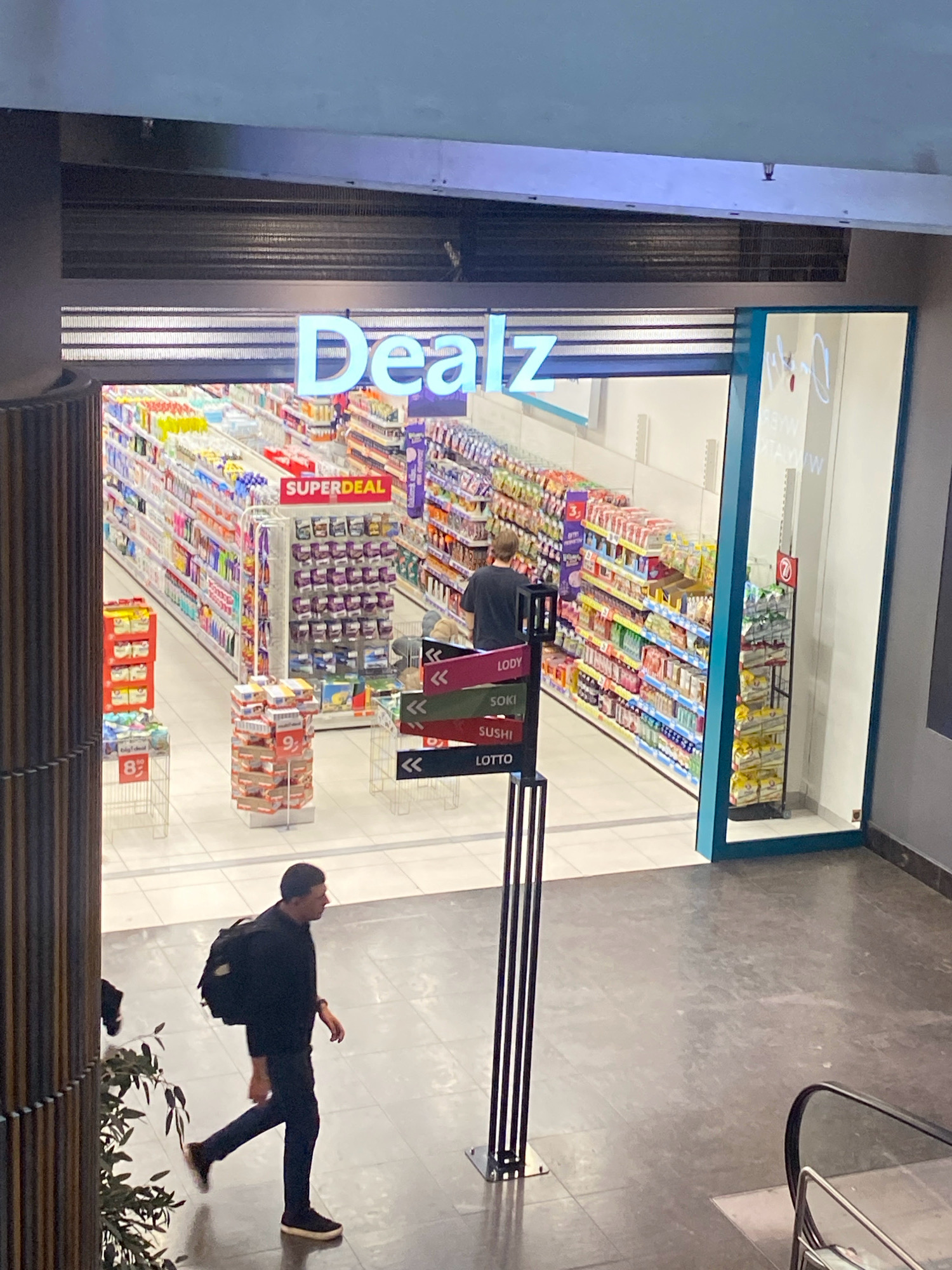 The image shows the interior of a "Dealz" store, a discount retailer, in Nowy Sącz, Poland. The store is brightly lit and stocked with various merchandise on shelves. The entrance to the store features large, light blue lettering spelling out "Dealz."
Inside, a person wearing a dark t-shirt and pants is browsing the aisles. They are facing away from the camera. Several aisles of products are visible, including what appear to be food items, household goods, and possibly toiletries. Signs indicating "SUPERDEAL" are visible on some displays.
In the foreground, a man wearing a black long-sleeved shirt, dark pants, and a black backpack is walking away from the camera, towards the left side of the frame. He is walking on a tiled floor. To the right of the man, part of an escalator can be seen. There are also some black vertical stands with directional arrows and text labels, such as "LODY" (ice cream), "SOKI" (juices), "SUSHI," and "LOTTO," suggesting these are categories or promotions within the store or related services. Some green foliage is visible on the left side of the image.