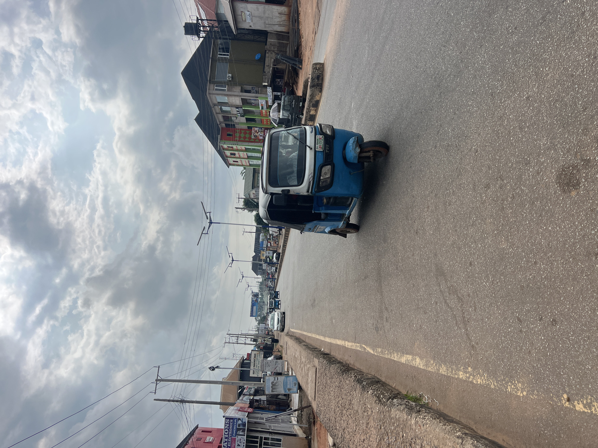 In Okpanam, Nigeria, a blue and white auto-rickshaw is captured in motion on a paved road, traveling away from the viewer and slightly to the right. The auto-rickshaw features a white canopy, a dark blue front section, and a visible license plate. The road is lined with a raised concrete curb on the left side, which is worn and shows signs of age, with sparse vegetation growing through cracks.
To the left of the road, several businesses are situated, including a building with a prominent pink facade displaying signage for "ATIONS ELECTRONICS" and "NOKIA." Adjacent to this, other shops and establishments line the street, with various signs and awnings visible. Further down the road, a white sedan is seen, and in the distance, multiple other vehicles and more auto-rickshaws are present, indicating moderate traffic.
The buildings on the right side of the road are a mix of modern and older structures. One building has a dark roof and a facade with green and red accents, featuring multiple windows. Another building to its right appears to be under construction or has an unfinished appearance, with exposed materials. Overhead, power lines crisscross the sky, supported by utility poles, and streetlights are spaced along the road.
The sky is overcast with dramatic, textured clouds, suggesting a day with variable weather conditions. The overall scene depicts a typical street in a Nigerian city, characterized by commercial activity, transportation, and the infrastructure common to such an environment. The lighting is diffused due to the cloud cover, casting a soft light on the scene.