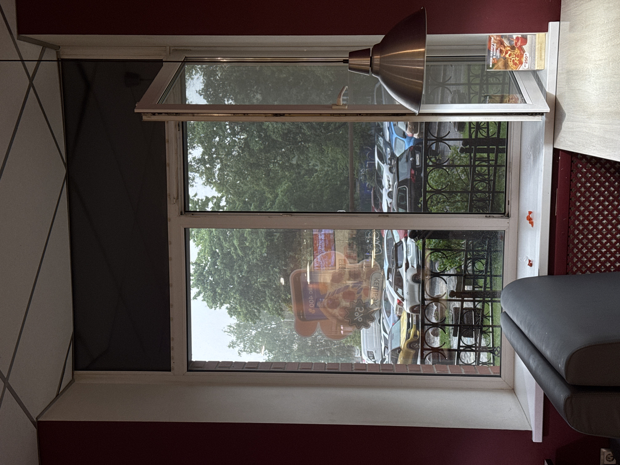 The image shows a view through a window from an indoor setting, likely a restaurant, looking out onto a street with parked cars and trees. The window is partially open. Inside, a metallic lampshade hangs from the ceiling. On the windowsill, there is a small advertisement card for a pizza. The walls are painted a deep red. Outside, it appears to be daytime, with an overcast sky. The parked cars suggest an urban or suburban environment. There are no people visible in the image.