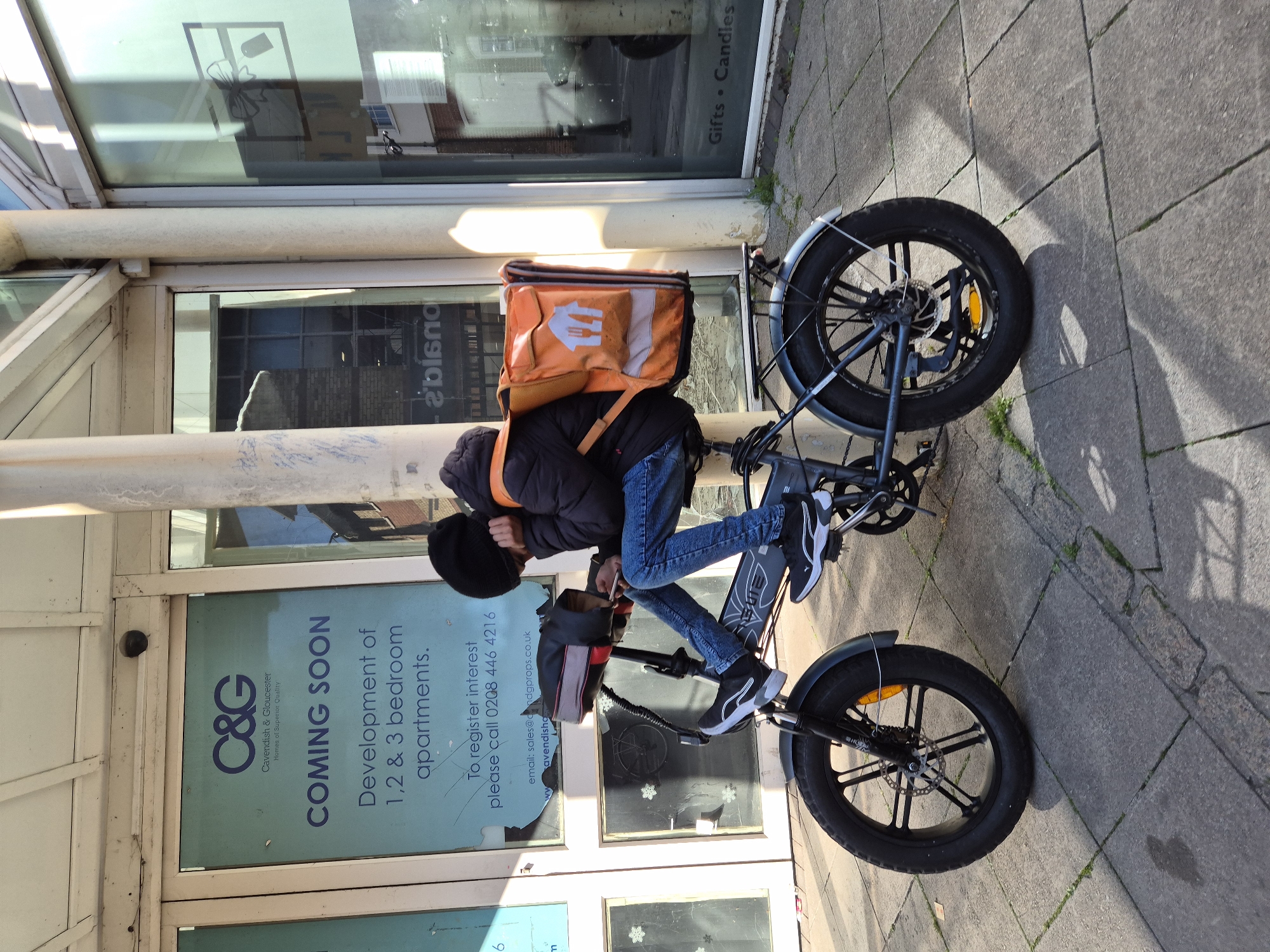 The image depicts a food delivery rider taking a break on an urban sidewalk, likely in West Suffolk, United Kingdom, given the context. The primary subject is a person, dressed in a black puffy jacket, blue jeans, a black beanie, and black and white Puma sneakers. They are leaning against a light-colored pillar or pole, with their head lowered, suggesting they are resting or checking a device.
Strapped to their back is a prominent orange insulated delivery bag featuring a white logo of a stylized house with cutlery, characteristic of food delivery services such as Just Eat or Foodpanda. Next to the person is a dark-colored electric bicycle, equipped with distinctive fat tires, disc brakes, and a rear rack. A small, dark-colored bag is also attached to the bicycle's handlebars.
The scene unfolds on a paved sidewalk, made of grey rectangular blocks, alongside a modern building with large glass windows. The bright conditions of a sunny day are evident from the sharp shadows cast by the bicycle and the person onto the pavement.
Reflected in and displayed on the building's windows are various signs. A large blue and white sign announces a development: "C&G Cavendish & Gloucester Homes of Superior Quality", "COMING SOON Development of 1,2 & 3 bedroom apartments. To register interest please call 0208 446 4216" and an email address "sales@candgprops.co.uk cavendishg". Another reflection or sign partially visible reads "Gifts • Candles". The overall setting suggests a commercial or residential area within a town or city.