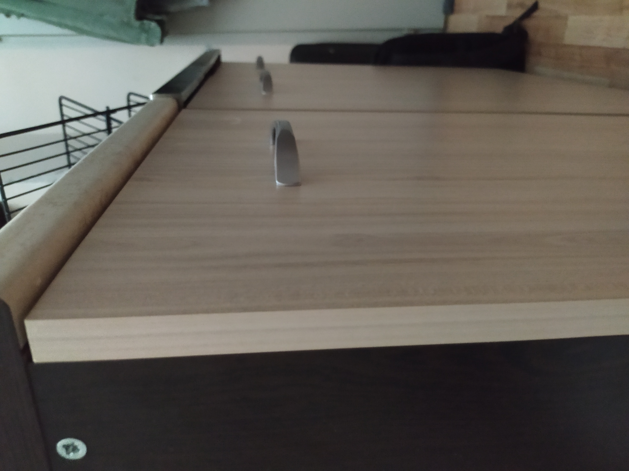 The image shows the top of a cabinet or dresser. The top surface is a light wood-grain laminate, and the front edge has a thicker band of the same material. Below the top surface is a darker, possibly brown or black, material. There are three silver-colored drawer pulls visible on the top surface. A metal wire rack is visible on the left side. In the background, there is a black object, possibly a bag, and what appears to be a wall. A faint greenish object is visible at the very top of the frame on the left. The image shows no people or location.