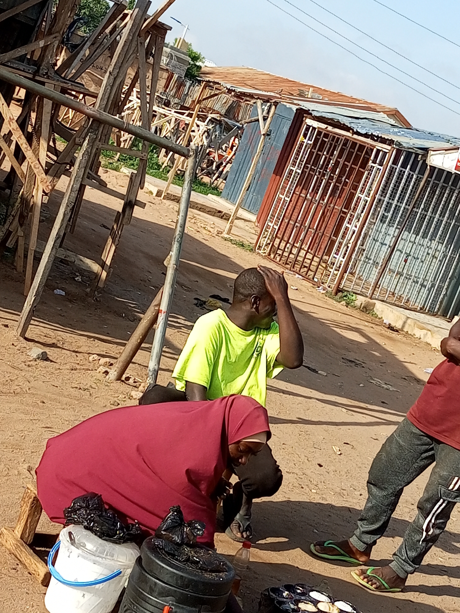 In Tempure, Nigeria, an outdoor scene depicts several individuals near makeshift structures and market stalls. In the foreground, a person wearing a red headscarf and garment is crouched down, interacting with two buckets, one white and one black, both containing black plastic bags. To the right, a person in a maroon t-shirt and patterned grey pants is standing, their feet visible in green flip-flops. Further back, another person in a bright lime green t-shirt is seated or crouching, holding their hand to their head. The ground is sandy and uneven, with scattered debris. In the background, a collection of wooden structures and corrugated metal market stalls are visible, suggesting a busy, perhaps informal, commercial area. The sky is a clear blue.