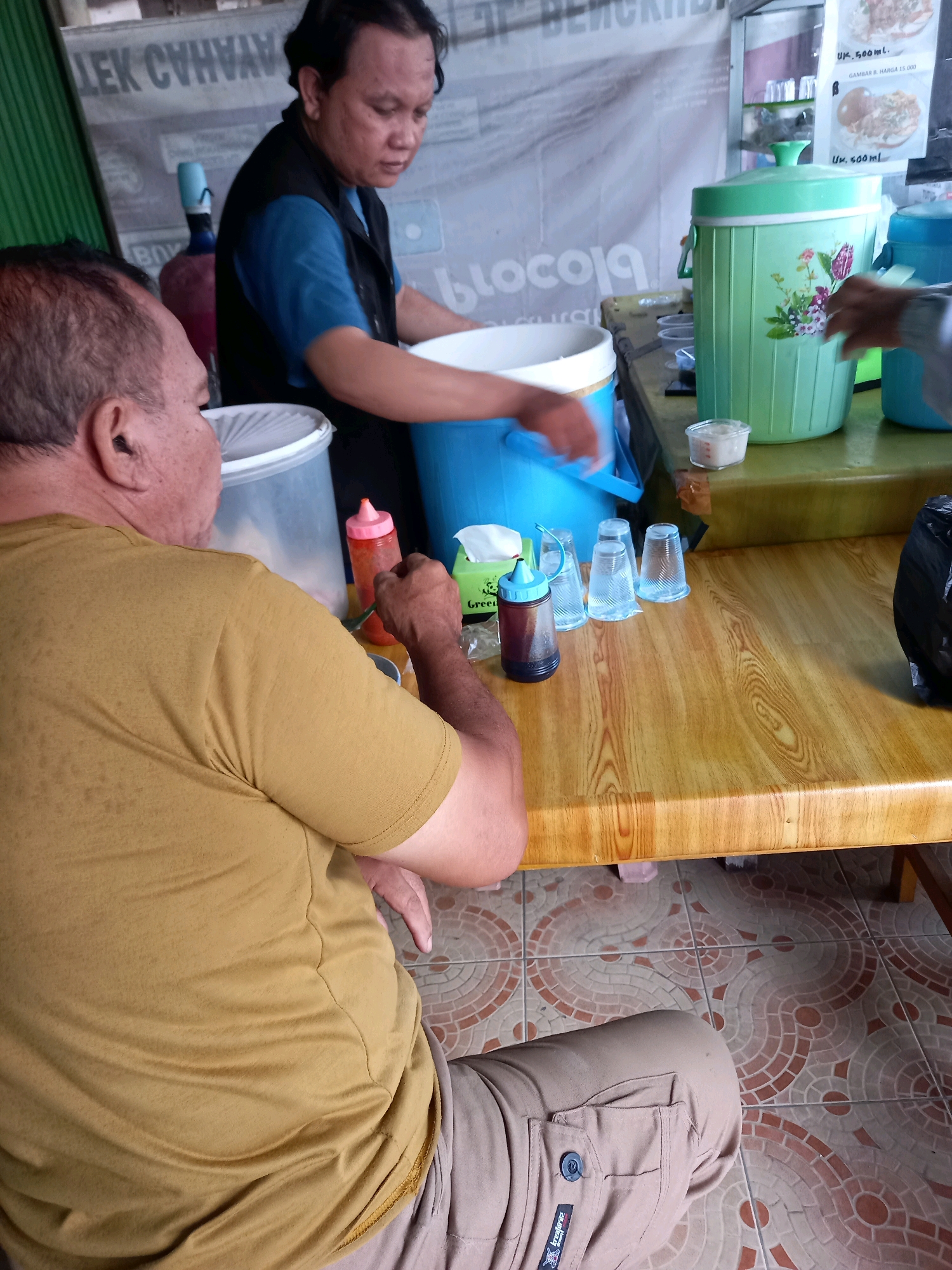 The image captures an indoor scene, likely a food stall or eatery in Samarinda, Indonesia. Two men are present, seated at a wooden table. One man, viewed from the back, wears a mustard-yellow t-shirt and light brown pants. He appears to be eating or preparing to eat, with his right arm extended towards the table. The other man, standing and facing away from the camera, is wearing a blue shirt and a dark vest. He is attending to a large blue bucket.
On the table, several items are visible: a pink condiment bottle, a bottle of dark liquid with a blue cap, a stack of clear plastic cups, a white tissue box, and a small container with white contents. A large green beverage dispenser with floral decorations stands prominently on a higher surface behind the table. The background features signage with Indonesian text. The floor is tiled with a geometric pattern. The overall impression is of a casual, utilitarian setting where food and drinks are served.
