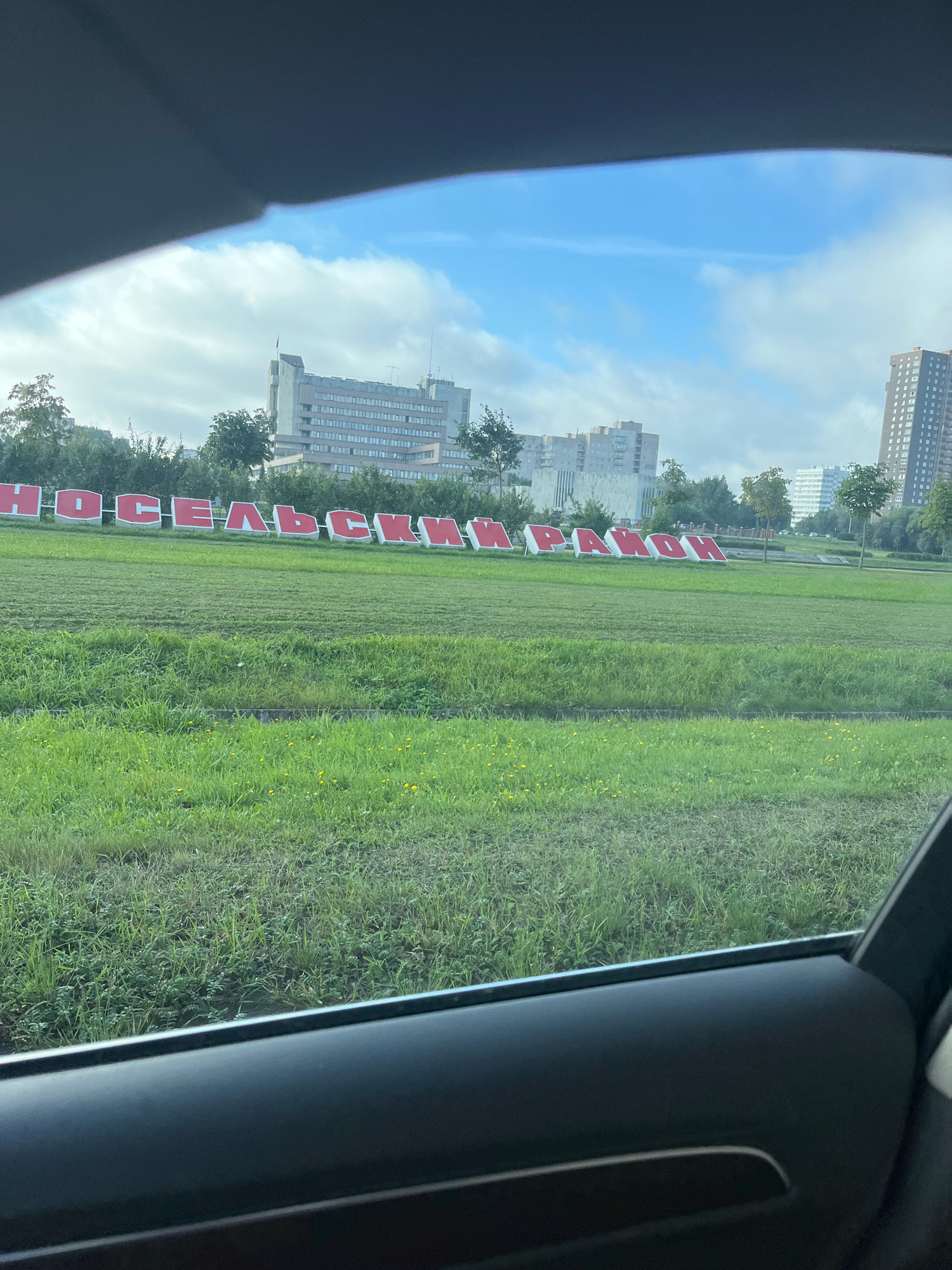 The image shows a large, red-lettered sign spelling out "ХОСЕЛЬСКИЙ РАЙОН" (Khoselsky District) in front of several multi-story buildings under a cloudy sky. The sign is situated on a well-maintained grassy area, possibly a park or roadside verge. The foreground shows the interior of a vehicle, indicating the photo was taken from a car window. The architecture of the buildings suggests an urban environment, consistent with a district in Saint Petersburg, Russia. No people are visible in the image.