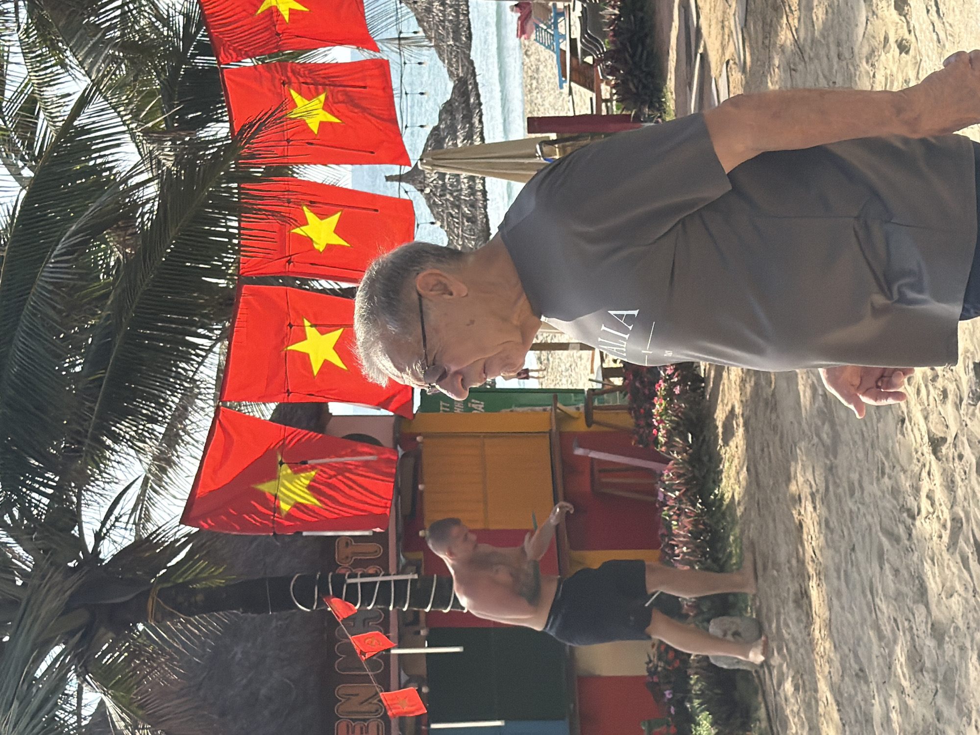 The image captures a beach scene in Đà Nẵng, Vietnam. Vietnamese flags hang vertically from a palm tree on the left. An older man wearing glasses and a gray shirt stands on a sandy beach facing to the left. A man with tattoos and wearing black shorts is walking on the sand in the distance. A beach is visible with some umbrellas in the background.