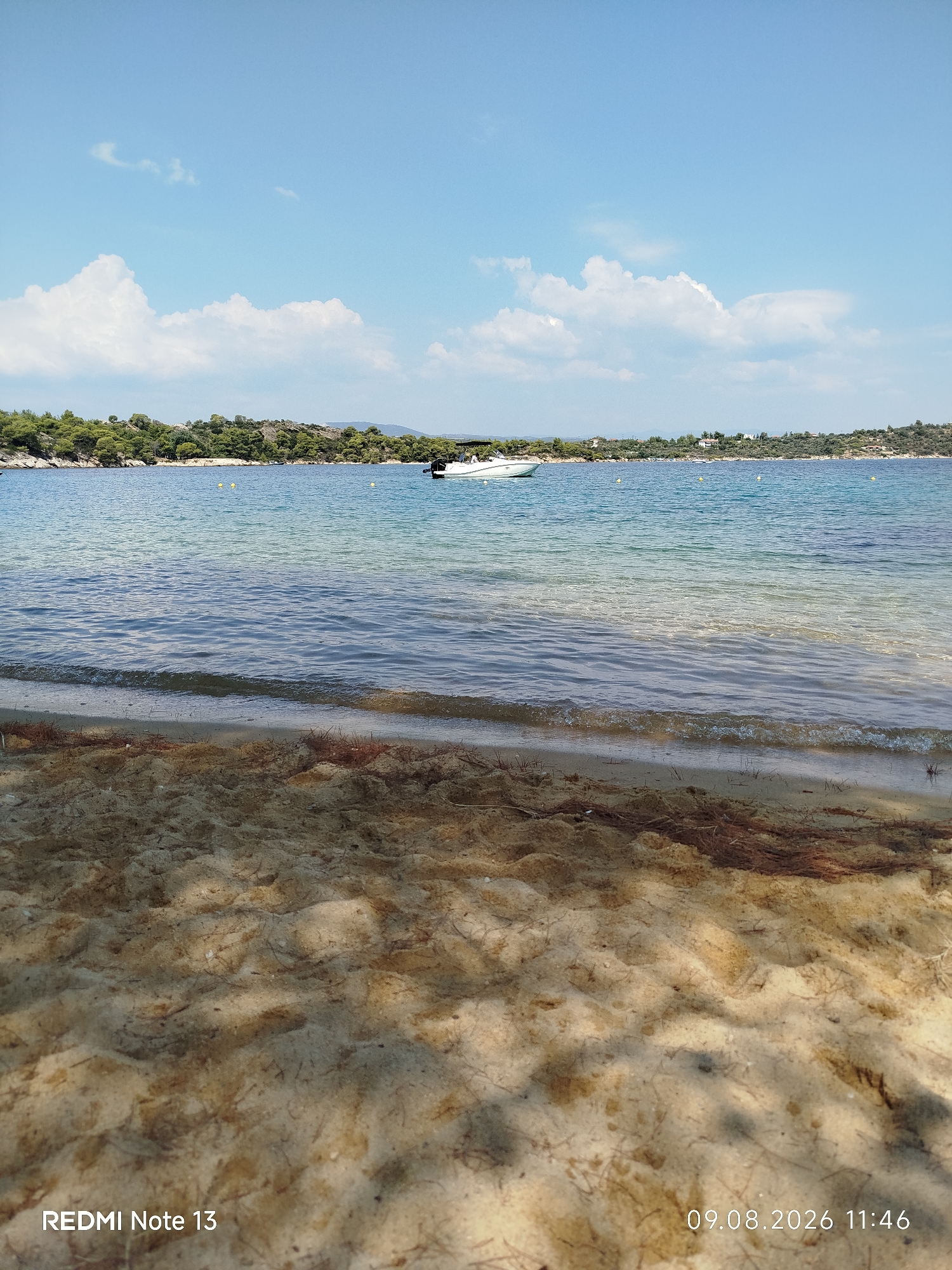 The image shows a serene coastal scene in Sithonia Municipal Unit, Greece, with a sandy beach in the foreground and the clear blue sea stretching towards a verdant, tree-covered coastline. A white motorboat is anchored in the tranquil waters, with a few yellow buoys visible in the distance. The sky is a vibrant blue, dotted with fluffy white clouds, suggesting a pleasant, sunny day. The sand on the beach is light brown and appears somewhat dry, with scattered pine needles and debris. The water is shallow and transparent near the shore, revealing the sandy seabed, and becomes a deeper blue further out. The overall impression is one of a peaceful, idyllic summer day at the seaside.