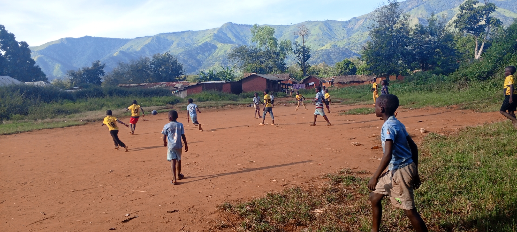 In the city of Ngalula, Democratic Republic of the Congo, a group of boys play soccer on a dirt field. Mountains are visible in the background. Some boys wear yellow shirts with numbers on the back, while others wear blue. There are houses with thatched roofs near the edge of the field.
