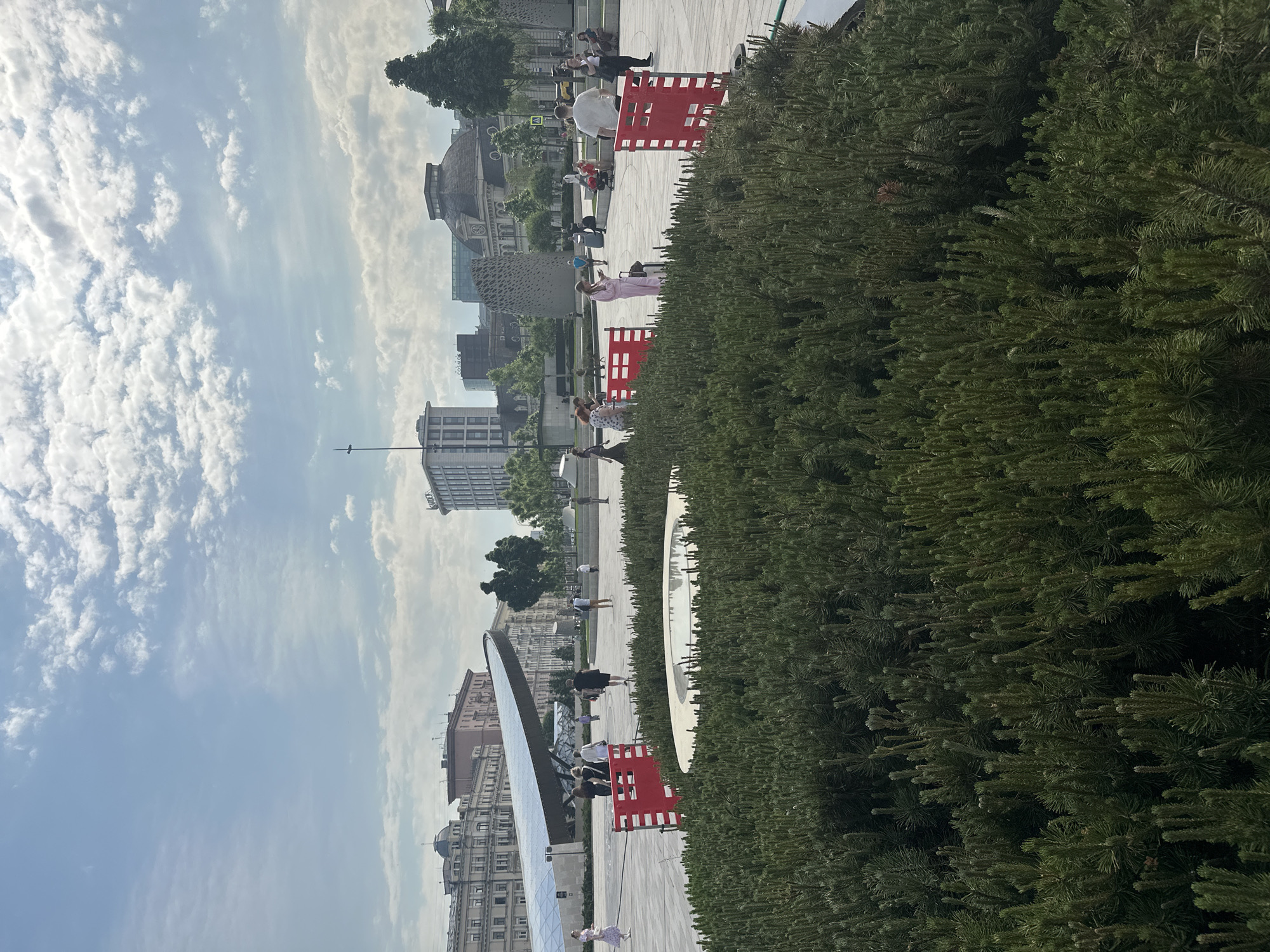 The image depicts a scene in Moscow, Russia, showcasing a public space with a mix of modern and classic architectural elements.
In the foreground, dense green shrubbery fills the right side of the frame, creating a natural boundary. A paved walkway extends into the distance. Scattered along the walkway are red and white barriers. Various individuals, including men and women of differing ages, are dispersed throughout the scene; they appear to be walking or standing, engaged in casual activities.
Buildings in the background exhibit a range of architectural styles, from traditional to contemporary. The sky overhead is partly cloudy.