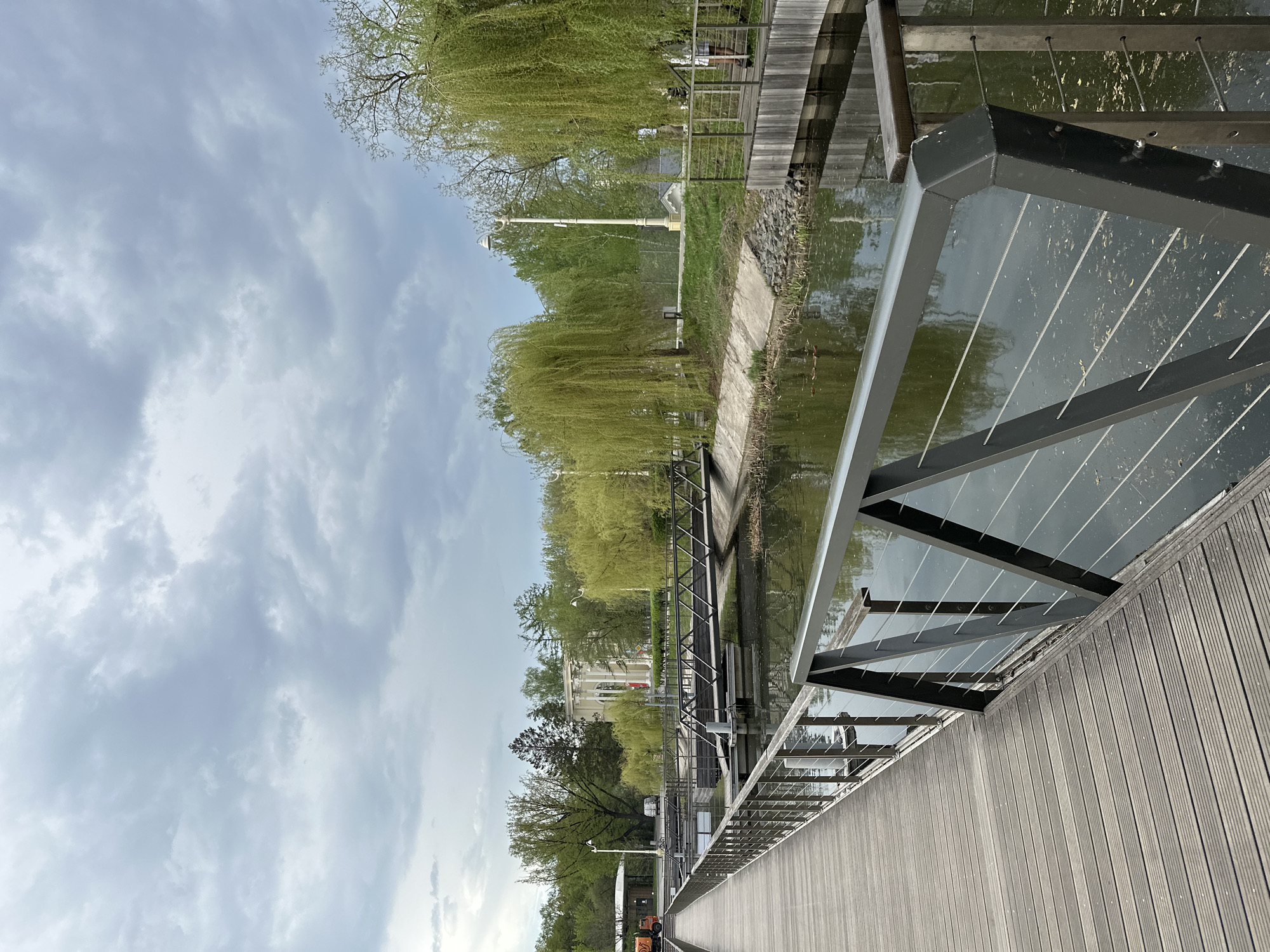 A partially obstructed view of a landscape is seen from a boardwalk. The boardwalk is made of grey wood. The boardwalk is lined with a steel railing. Behind the railing is a body of water. On the far side of the water is vegetation and a building. The sky is cloudy. Based on the scene's characteristics, it could be located in the city of Moscow, Russia.