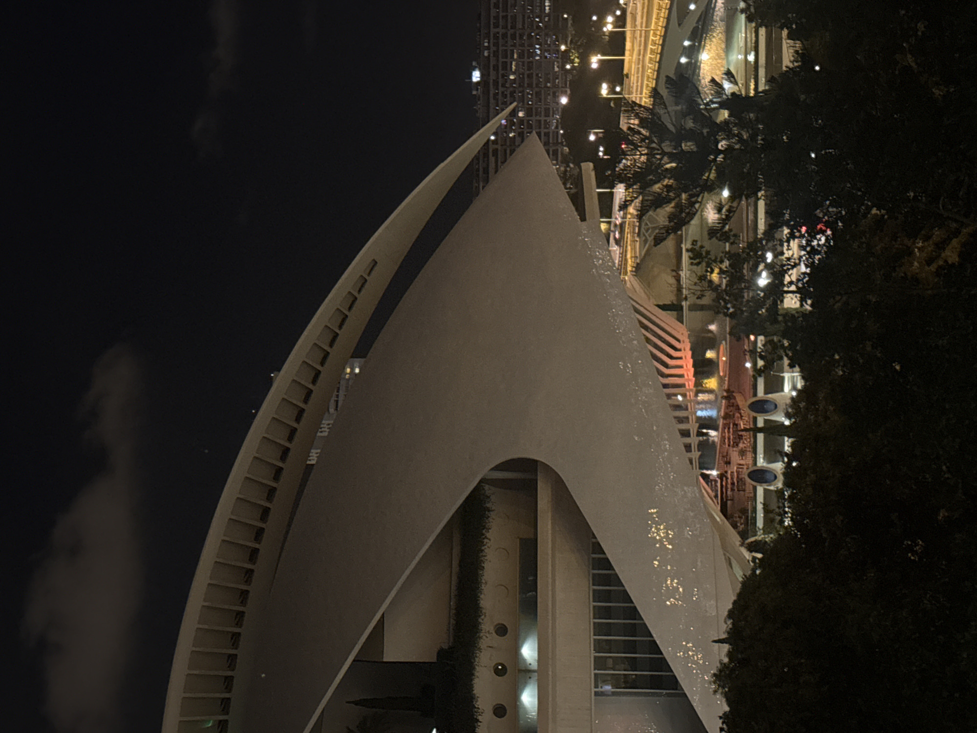 The image shows a night view of the City of Arts and Sciences in Valencia, Spain. The foreground features a large, curved, white architectural structure with a textured surface, possibly part of the Hemisfèric or the Oceanogràfic. Below this structure, there are glimpses of vegetation and illuminated circular windows or portholes. In the middle ground, a body of water reflects the lights from nearby structures, and various levels of illuminated pathways and bridges are visible, with cars driving on them. Tall, modern buildings with numerous illuminated windows are seen in the background. Silhouetted trees and foliage are present in the very foreground, obscuring some of the lower parts of the scene. The sky is dark and cloudy.