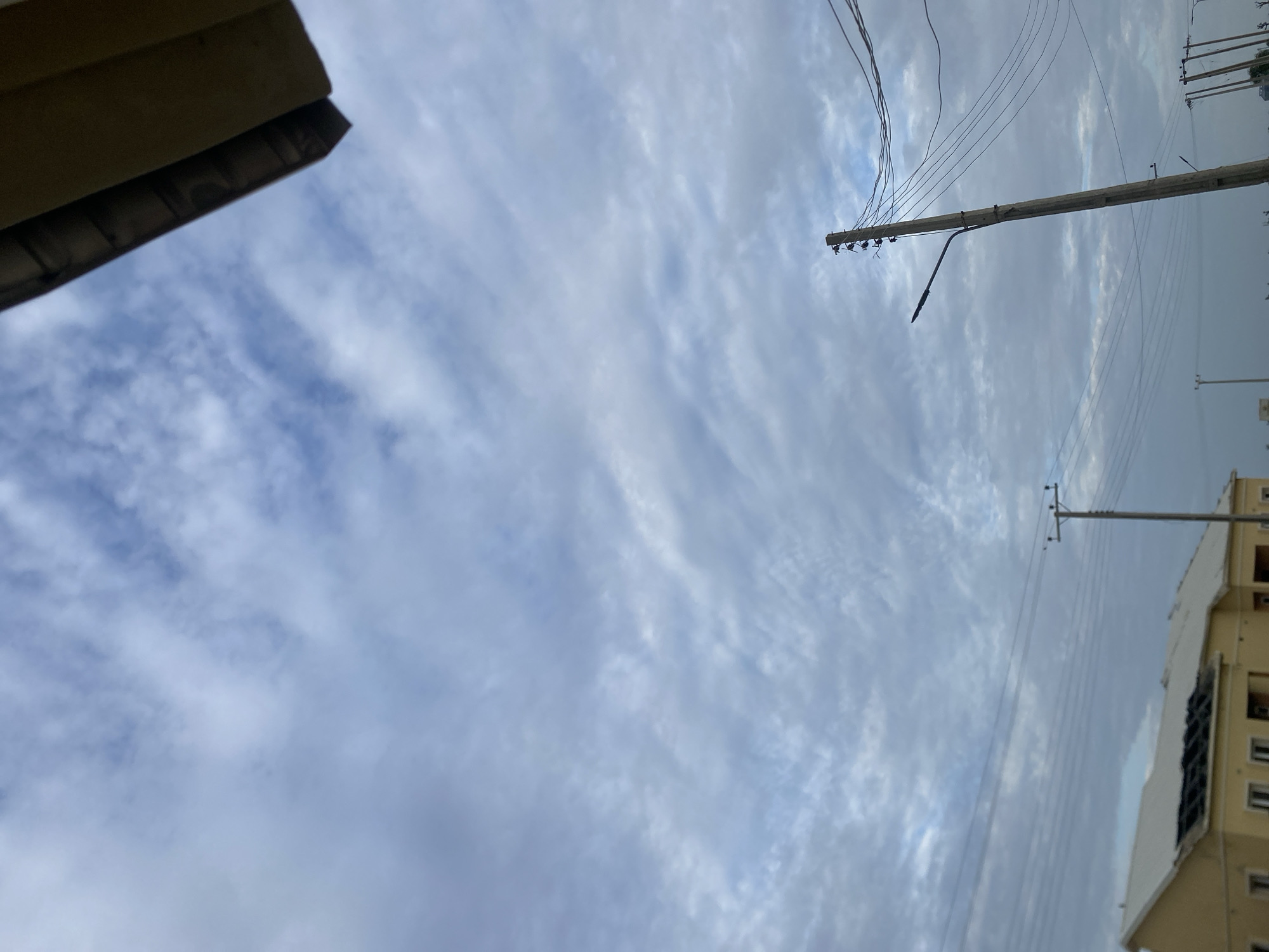 The image depicts an outdoor scene under a cloudy sky. In the lower left, a portion of a pale yellow, single-story building with a white roof is visible. The building appears to be under construction or renovation, with a section of the roof showing dark, open beams. Several utility poles with power lines crisscross the sky above the building. One prominent wooden utility pole stands in the mid-right of the frame, carrying numerous wires and a street lamp. Another utility pole with a street lamp is visible further in the background on the right. The sky is filled with a textured layer of grey and white clouds, suggesting an overcast or possibly approaching rainy weather. The scene appears to be in a semi-urban or developing area, consistent with a location in Dumnga, Nigeria. No people or animals are present in the image.