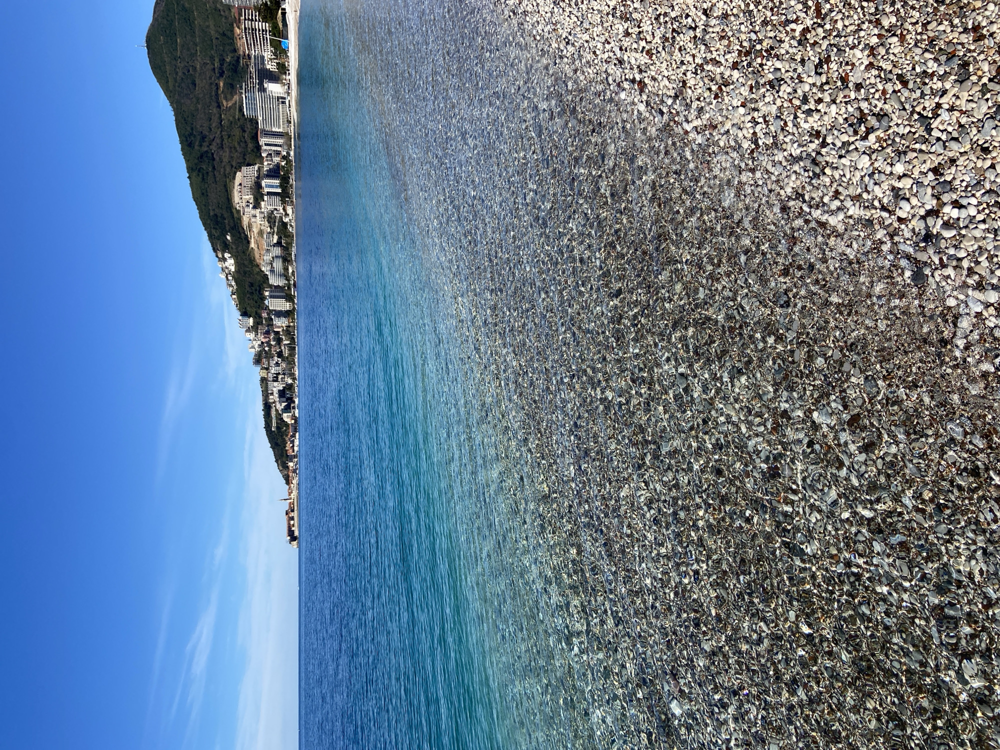 This photo captures a serene scene at Mogren Beach in Budva, Montenegro. The image is split between the azure sky and Adriatic Sea. The left side features a mountain with buildings lining its base, extending into the sea. The water transitions from deep blue to a crystal-clear expanse with the seabed visible, revealing pebbles. The right side of the image showcases the shore covered in small, light-colored pebbles. There are no people visible in the scene.