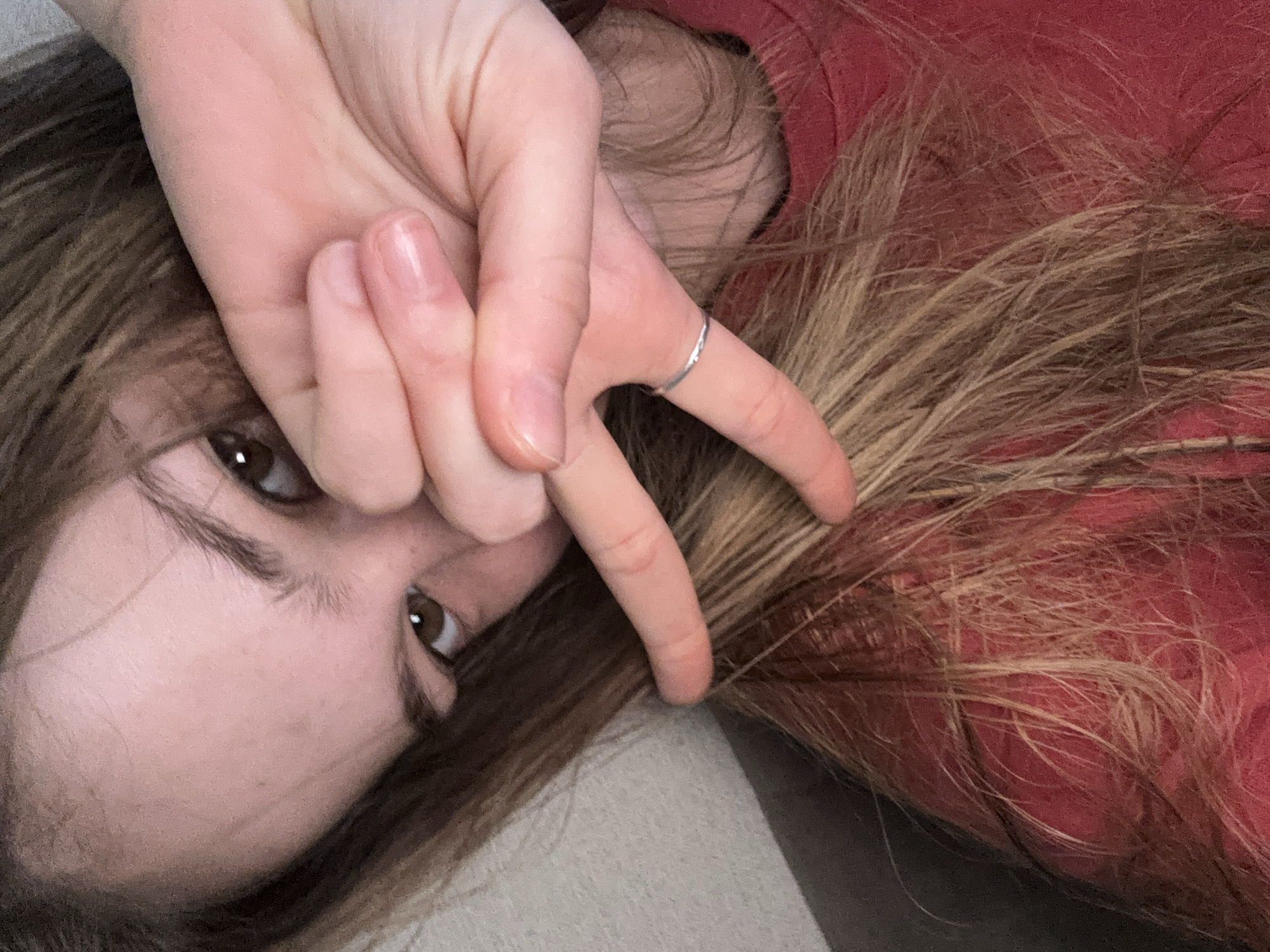 A close-up, high-angle shot shows a young person with long, brown hair covering part of their face. They are wearing a red shirt and have their hand up to their face, obscuring their eye. A thin silver ring is visible on their left ring finger. The background is a plain, light-colored surface. The image appears to be a selfie.