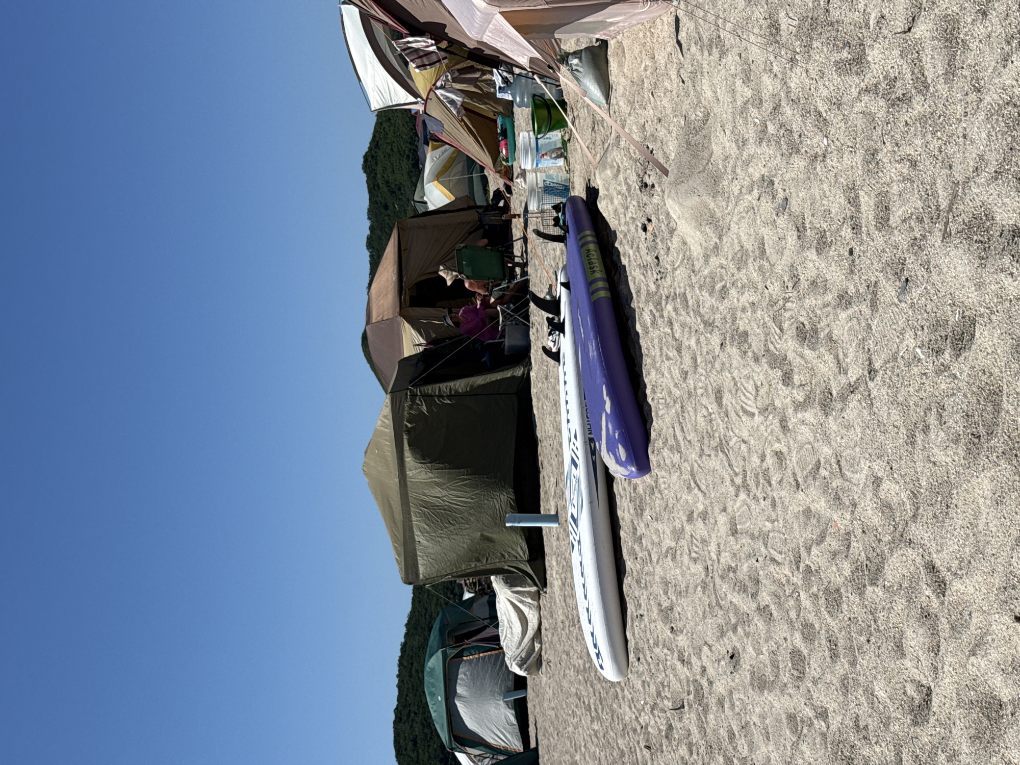 The image depicts a beach scene with several tents set up on the sand, suggesting a camping or outdoor event. Two surfboards, one white with blue accents and another purple, lie horizontally in the foreground. In the background, various tents of different colors and sizes are visible, with some individuals seated in chairs near one of the tents. The environment is characterized by a clear blue sky and sandy terrain, indicative of a coastal location. No specific details point to the city of Nakhodka, Russia, or its landmarks. The scene appears to be an active outdoor recreational area.
