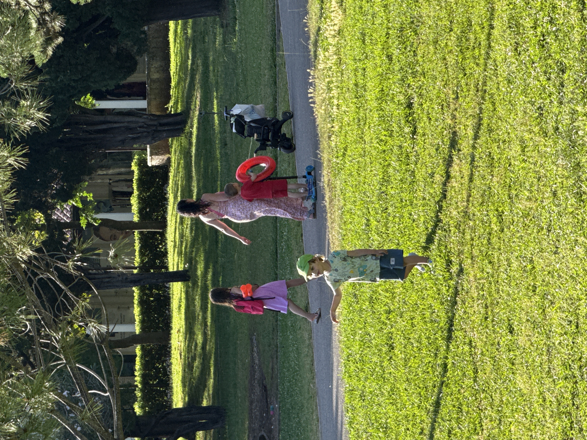 In Budva, Montenegro, a scene unfolds in a park-like setting. A woman in a long, floral-patterned dress walks alongside a young boy on a scooter, who holds a red inflatable ring. Beside them, a stroller is parked. Further ahead, a young girl in a lavender dress and a pink backpack walks on a paved path, holding orange water guns. In the foreground, a young boy wearing a green cap and a patterned shirt stands on the grass, pointing forward with his left arm. The environment is characterized by lush green grass, mature trees casting shadows, and a paved pathway, indicative of a public outdoor space.
