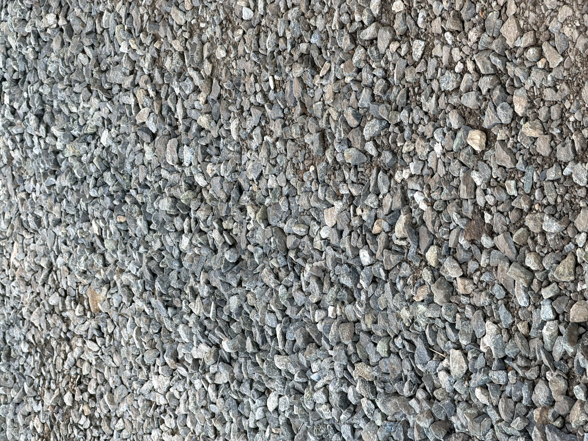 This image is a close-up, top-down view of a surface covered in crushed stone or gravel. The stones are predominantly gray with some variations in shade and texture, including lighter, almost white pieces and darker, almost black ones. Many of the stones are angular and irregular in shape, typical of crushed rock. The gravel appears to be loosely spread, with some areas showing small amounts of dirt or fine sediment interspersed among the larger pieces. The lighting suggests it is daytime, possibly outdoors, as there are no artificial light sources visible and the illumination appears natural. There are no people or specific objects, beyond the gravel itself, that are the focus of the image. The texture and composition of the gravel suggest a ground surface, possibly a path, driveway, or construction site in Bol'shaya Ven'ya, Russia. There is no discernible text in the image.