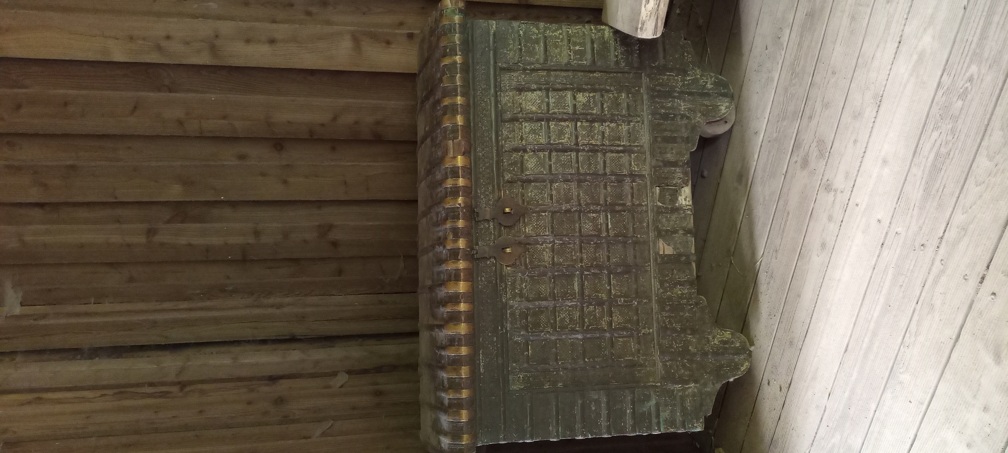 An aged, dark green wooden chest with metal accents sits on a light-colored wooden floor. The chest has decorative carvings and two metal clasps on the front. It is positioned against a vertical wooden plank wall. The overall scene is indoors, suggesting a rustic or historical setting. There are no people visible in the image. The location is not specified within the city of Gelsenkirchen, Germany.