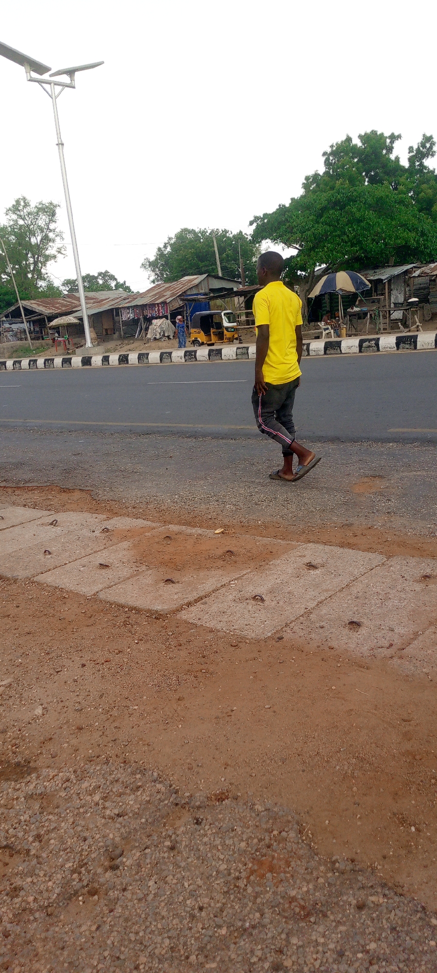 The image depicts a road scene in Zango-ndaji, Nigeria. A man wearing a yellow t-shirt and dark pants with a pink stripe is walking on the side of the road. A yellow tricycle is visible on the other side of the road near a small building with a corrugated metal roof and painted supports. Various objects are located on the street, including what appears to be a table and chairs and an umbrella. The environment is open with vegetation in the background. A tall light pole stands to the left of the road. The foreground shows a dirt and gravel shoulder next to a concrete edge.