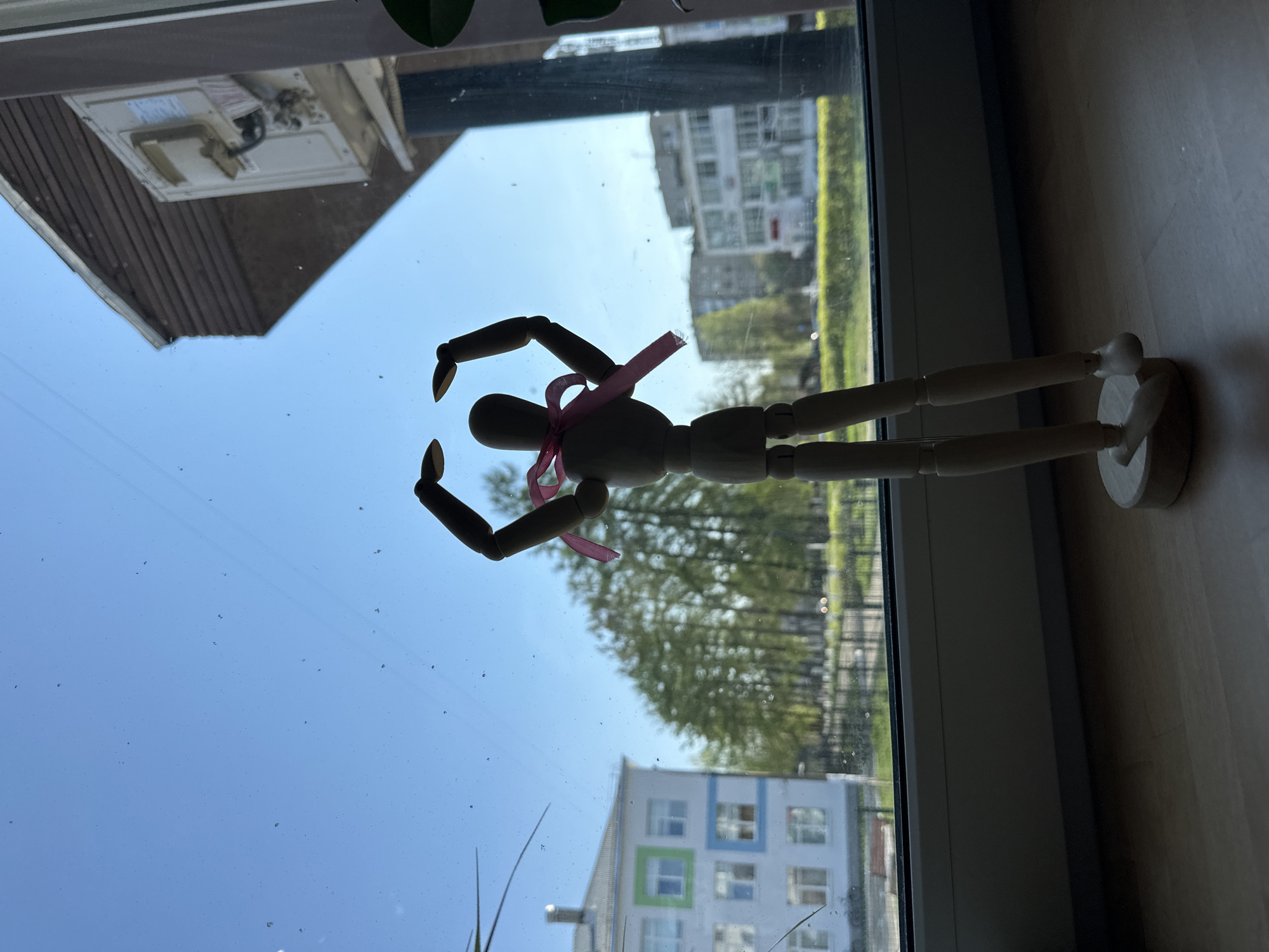 A wooden articulated mannequin figure with a pink ribbon tied around its torso stands on a windowsill inside a building in Chaikovsky, Russia. The mannequin faces the window, positioned with arms raised. The reflection in the window shows a building with a gable roof. Through the glass are views of other buildings, trees, and a clear blue sky, suggesting an outdoor setting with residential buildings. The windowsill appears to be made of wood.