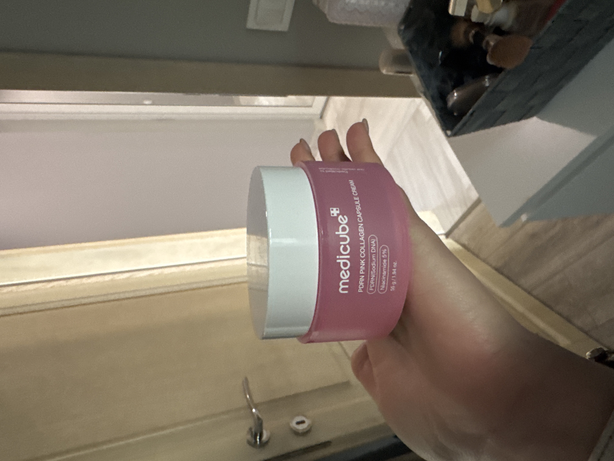 A hand holds a jar of "medicube PDRN PINK COLLAGEN CAPSULE CREAM". The jar is pink with a white lid, and the text on the label is legible, including the details "PDRN(Sodium DNA), Niacinamide 5%, 55 g/1.94 oz." The background shows a bathroom sink and faucet, and in the upper right corner, there is a partial view of a container with various items inside. Based on the style of the bathroom fixtures and the presence of the cream, the setting seems to be a personal residence, likely a bathroom or vanity area. Without additional specific location information, it's impossible to determine if the scene is located in Sofia, Bulgaria.