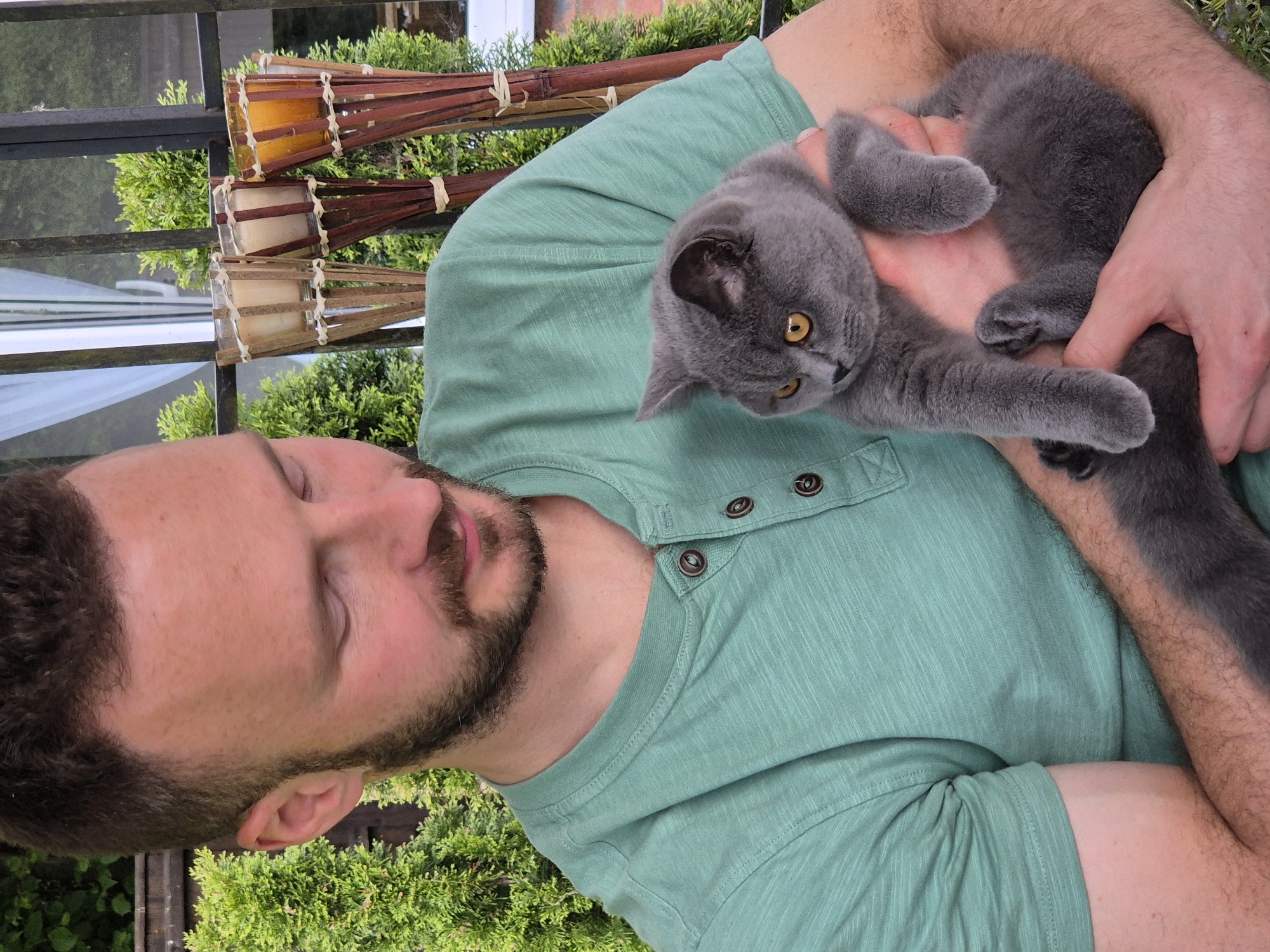 A light-skinned man with brown hair and a beard is holding a grey cat. The man is wearing a light green button-down shirt. The cat has yellow eyes and is looking to the left. In the background, there is greenery and a metal railing. There are also what appear to be lanterns above the railing. The location is West Suffolk, United Kingdom, although specific details within the city cannot be determined from this image.