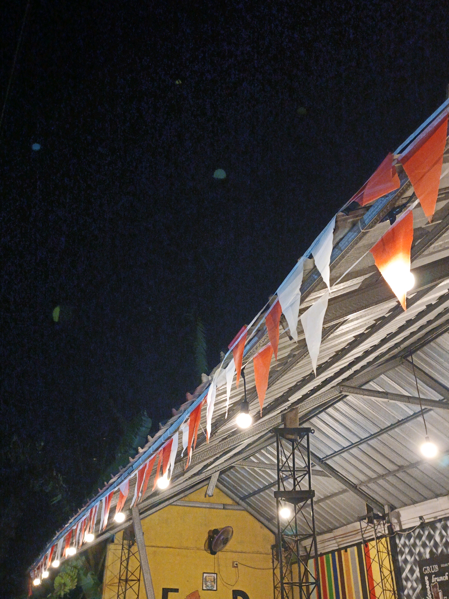 In Ketegan, Indonesia, an outdoor covered seating area features a string of red and white triangular flags draped along the roofline. The flags are illuminated by overhead lights, some of which are visible. The roof is made of corrugated metal, supported by a metal frame structure. Below the roof, a yellow building is visible, and a black fan is mounted on the wall. In the foreground, a sign with colorful vertical stripes is present, along with some text that is partially obscured. The background is a dark night sky with a few faint, blurry lights.