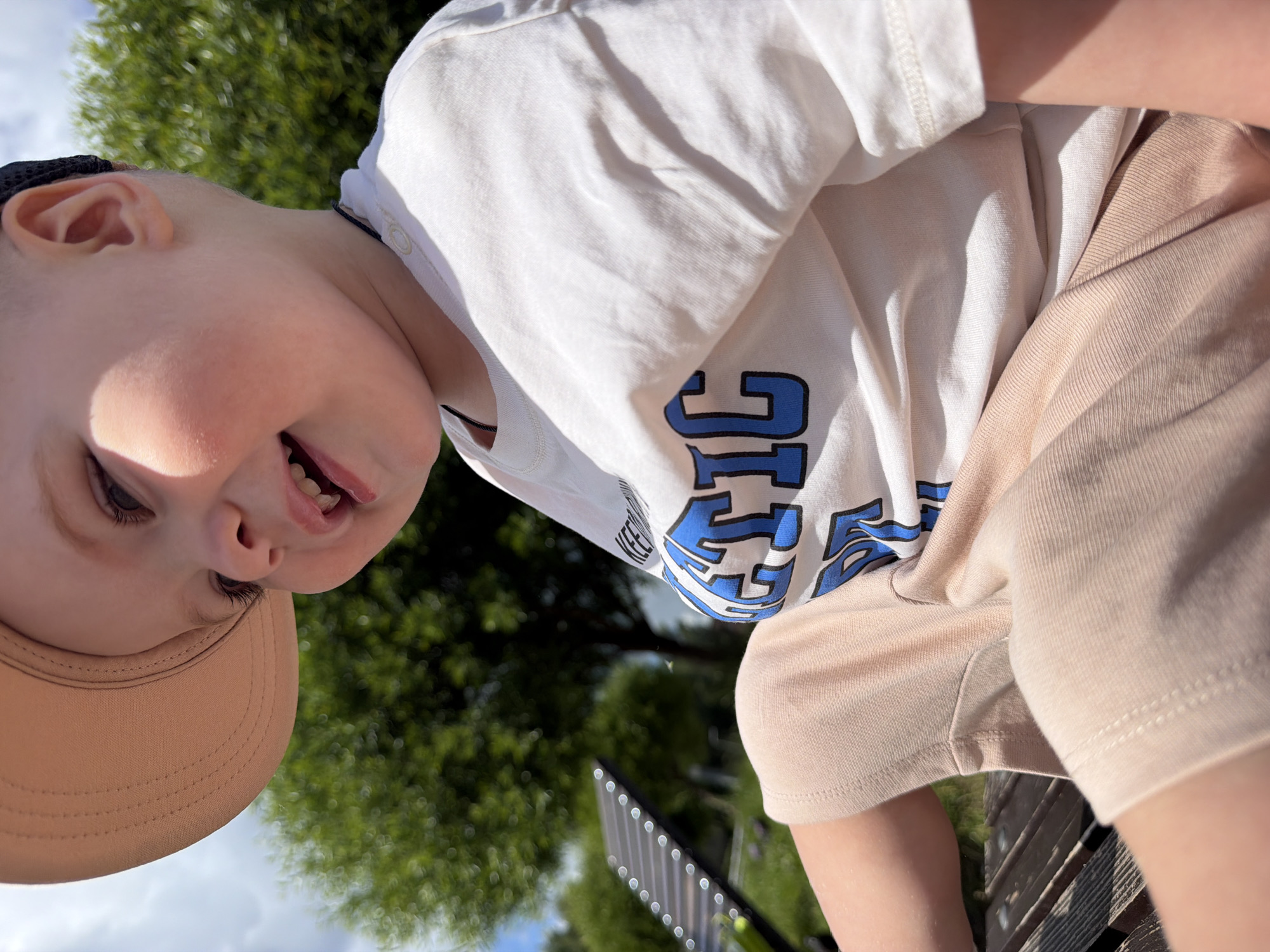 A low-angle, close-up shot shows a young child's face and torso outdoors. The child, wearing a white t-shirt with blue lettering that appears to say "KEEP GOING", and beige shorts, is looking down and smiling. A light-brown cap is visible on the child's head. The background is blurred, showing green foliage from trees and a bright sky, suggesting it is daytime with good weather. The child is likely sitting or being held, as evidenced by the perspective and the presence of what looks like a wooden bench below. The scene appears to be a casual outdoor setting, possibly a park or backyard, in Votkinsk, Russia.