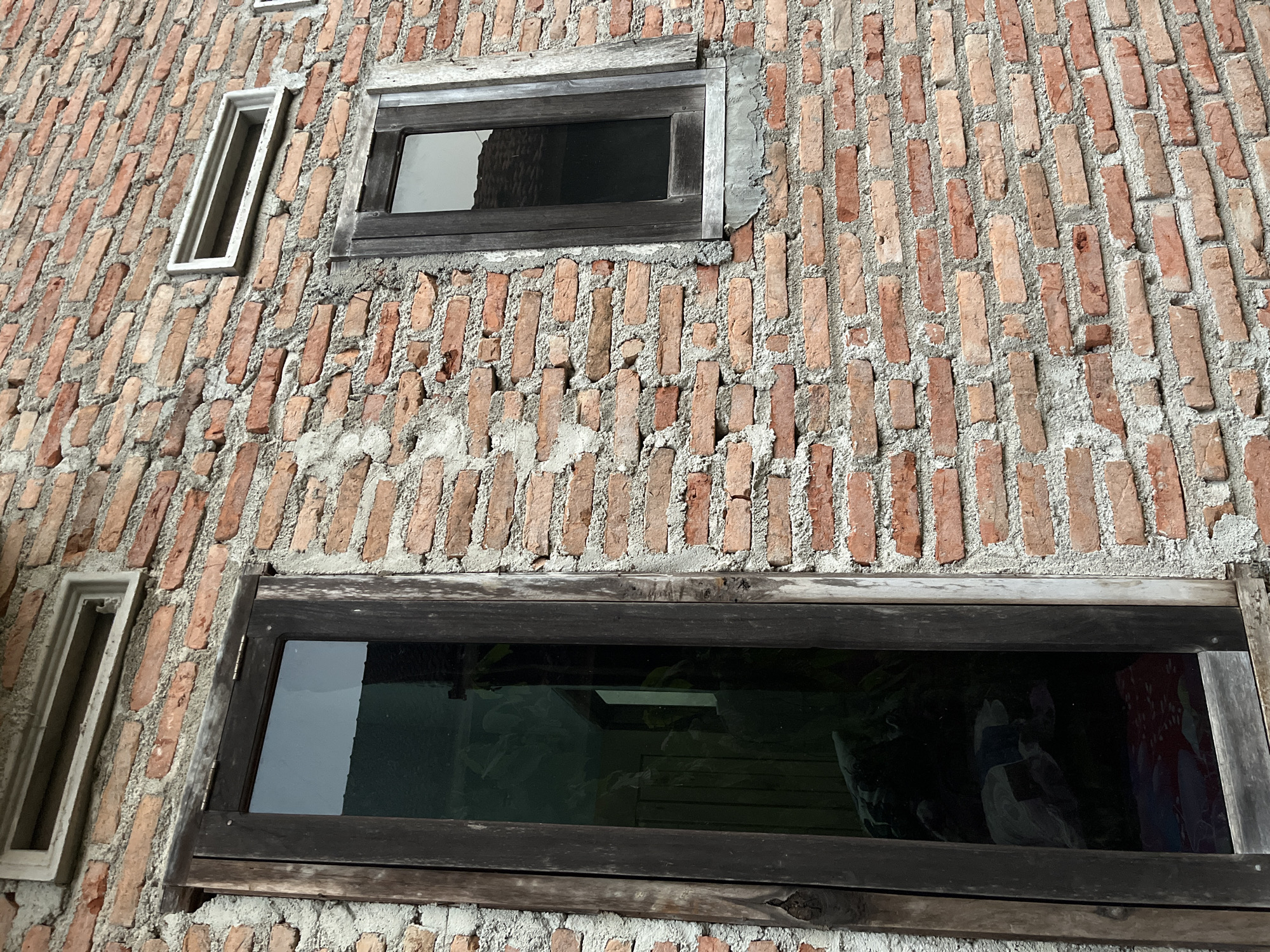 The image shows the exterior of a brick building in Kebumen, Indonesia, with two windows and a small vent. The wall is constructed of rough, reddish-brown bricks with visible mortar. The larger window on the left is tall and narrow, with a dark wooden frame. Its glass is reflective, showing a dark interior with hints of furniture and patterned fabric. A smaller window on the right, also with a dark wooden frame, is square and reflects the sky or a rooftop. Above the larger window and to the right of the smaller window are rectangular concrete vents. The overall scene depicts a rustic architectural style, likely residential, with an unfinished or weathered appearance.