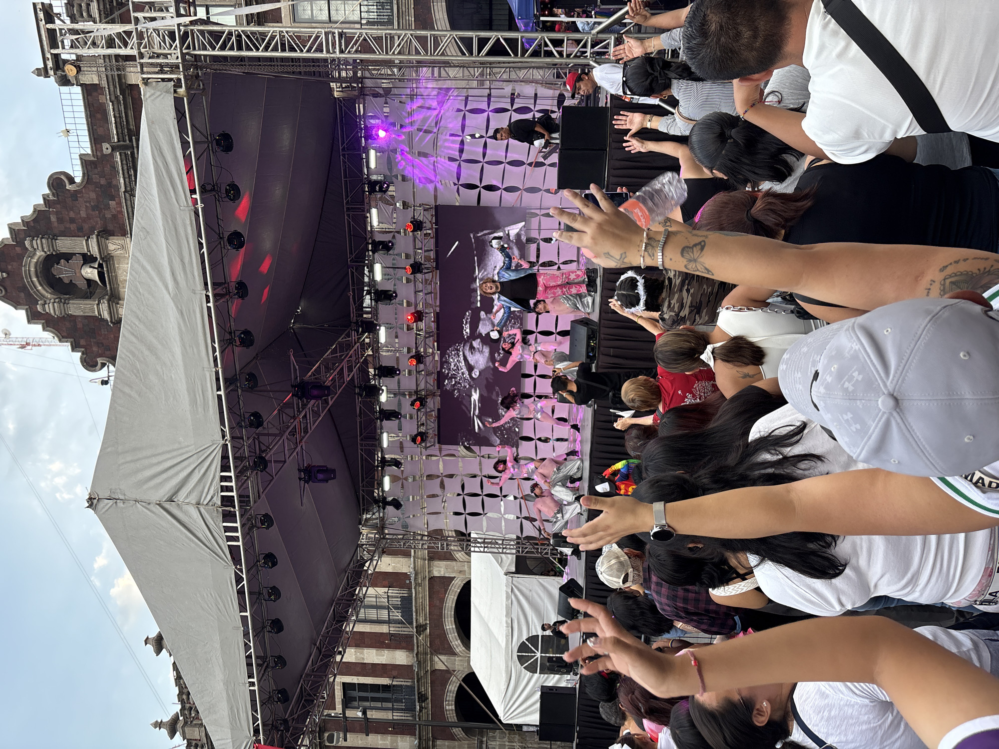 The image depicts an outdoor performance or event taking place in the Zocalo of Cuernavaca, Mexico. A stage is set up with a large screen displaying images, and several performers are on stage, some dancing in metallic outfits and others interacting with the audience. A man in a blue jacket and pink pants is front and center, appearing to be the main performer, with a DJ or sound technician to his right. A crowd of people is gathered in front of the stage, many with their hands raised in the air, indicating engagement with the performance. The background shows colonial-era architecture characteristic of Mexican city centers. The event appears to be a public gathering with a festive atmosphere, possibly a concert or festival, given the stage setup and the size of the crowd.