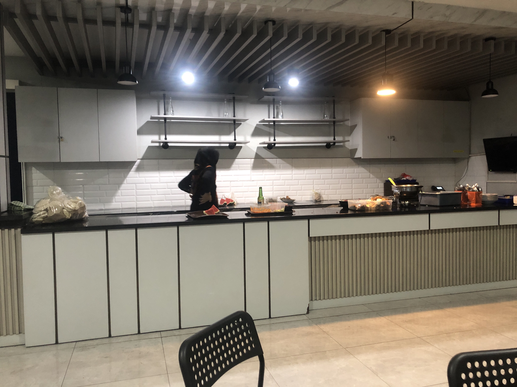 The image depicts a modern kitchen or pantry area. In the foreground, a black perforated chair is visible. The middle ground features a long countertop with various food items and containers, including a bag of snacks, slices of watermelon on a plate, a bowl of salad, and a bottle of green liquid. A person wearing a dark outfit and head covering is standing behind the counter, appearing to prepare or arrange food. The background includes white subway tile walls, white cabinets mounted above the counter, and open shelving units with glass bottles. The ceiling has a distinctive slatted design with modern pendant lights. The floor is tiled. The scene suggests a communal eating or food preparation space, possibly in an office or educational institution. The location is identified as Wonojati, Indonesia.