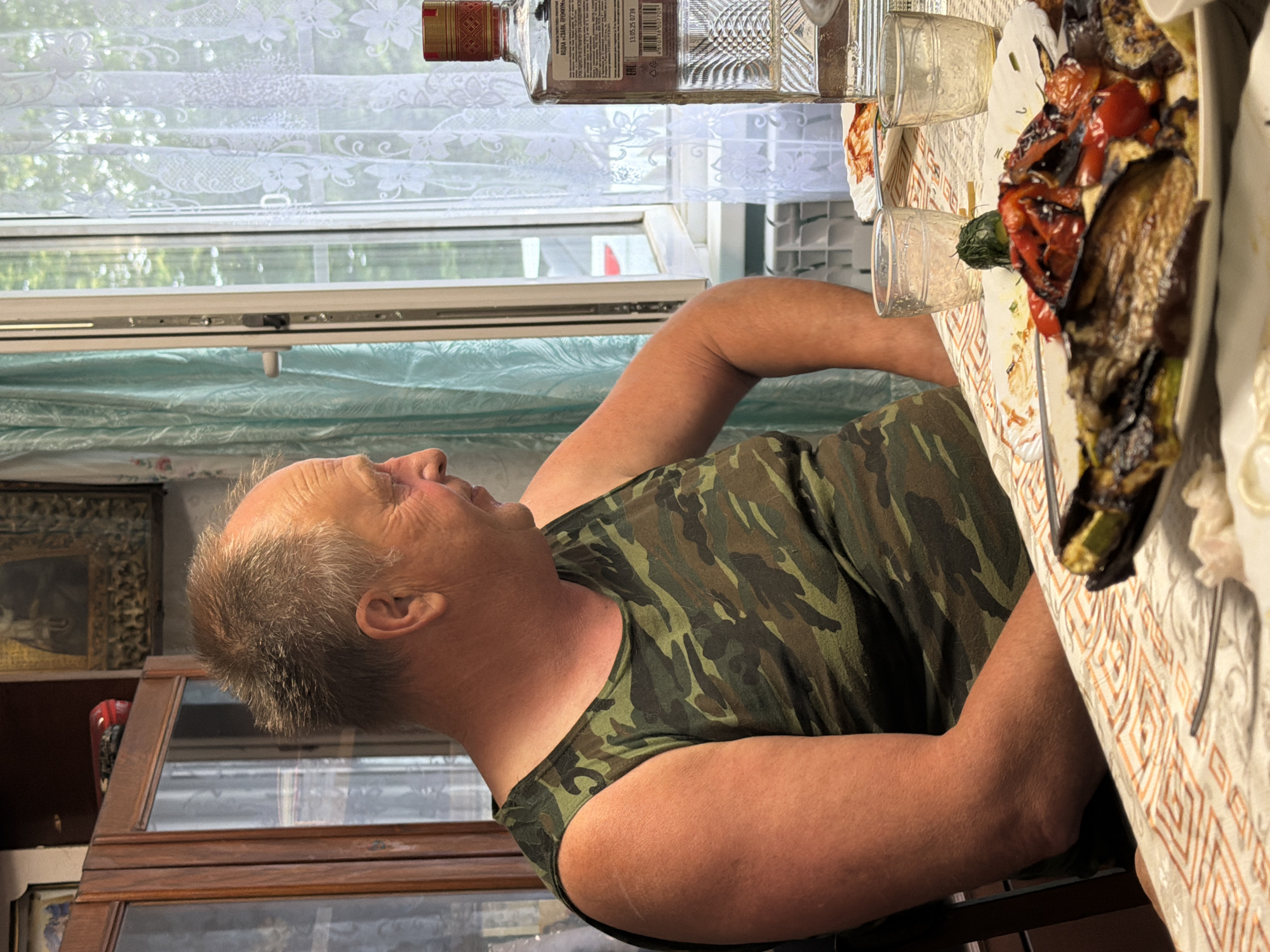 A man wearing a camouflage tank top is sitting at a table, leaning forward. He has fair skin and graying hair. The table is set with a meal that includes grilled vegetables such as eggplant and tomatoes. Two glasses, one partially filled with a clear liquid, are also on the table, along with a bottle of alcohol with a red and gold label. The background shows a window with white lace curtains, suggesting an indoor setting. Outside the window, trees and greenery are visible, indicating daylight. To the left of the man, a wooden cabinet with glass doors is partially visible, containing framed pictures and decorative items. The overall scene appears to be a casual meal at home, possibly in a rural or suburban area of Бронница, Russia. The lighting suggests it is daytime, but the specific time of day is not evident. The man's expression is neutral. The label on the alcohol bottle contains text in Cyrillic script, including what appears to be a date "13.05.25" and a time "07:39".