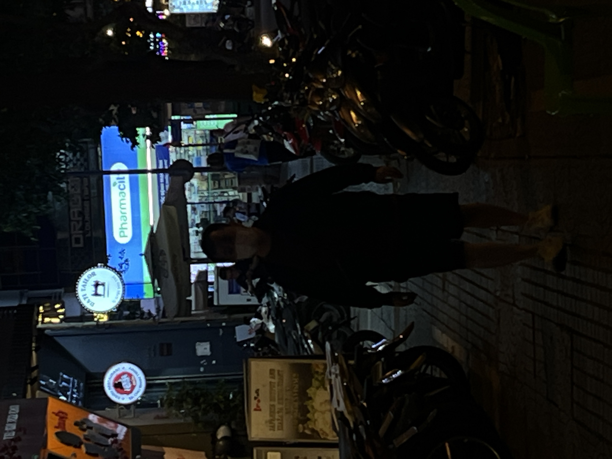 A man wearing a white face mask and a dark long-sleeved shirt and shorts walks down a sidewalk at night. The sidewalk is paved with patterned tiles and is lined with parked motorcycles. Behind the man, a bright blue sign with white text that reads "Pharmacil" is illuminated. To the left of this sign, a circular sign with "DAZI TAILOR" and a sewing machine graphic is visible. Further to the left, another circular sign with a red graphic is present. The environment suggests a commercial street in Thủ Đức, Vietnam, with multiple businesses indicated by the signs and the presence of neon lights and shop fronts in the background.